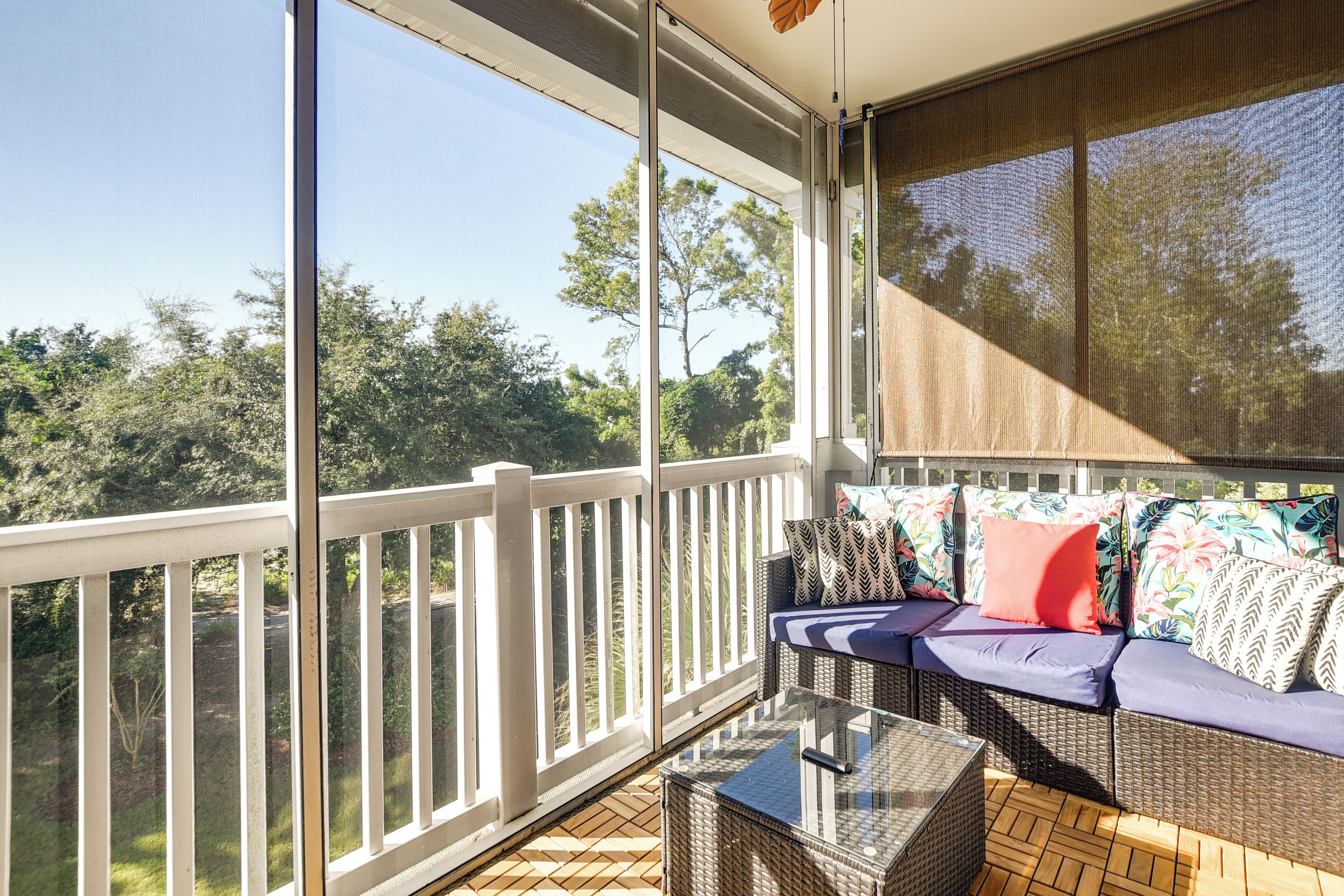 A covered balcony with a wicker sofa, a glass table, and a view of trees and sky.