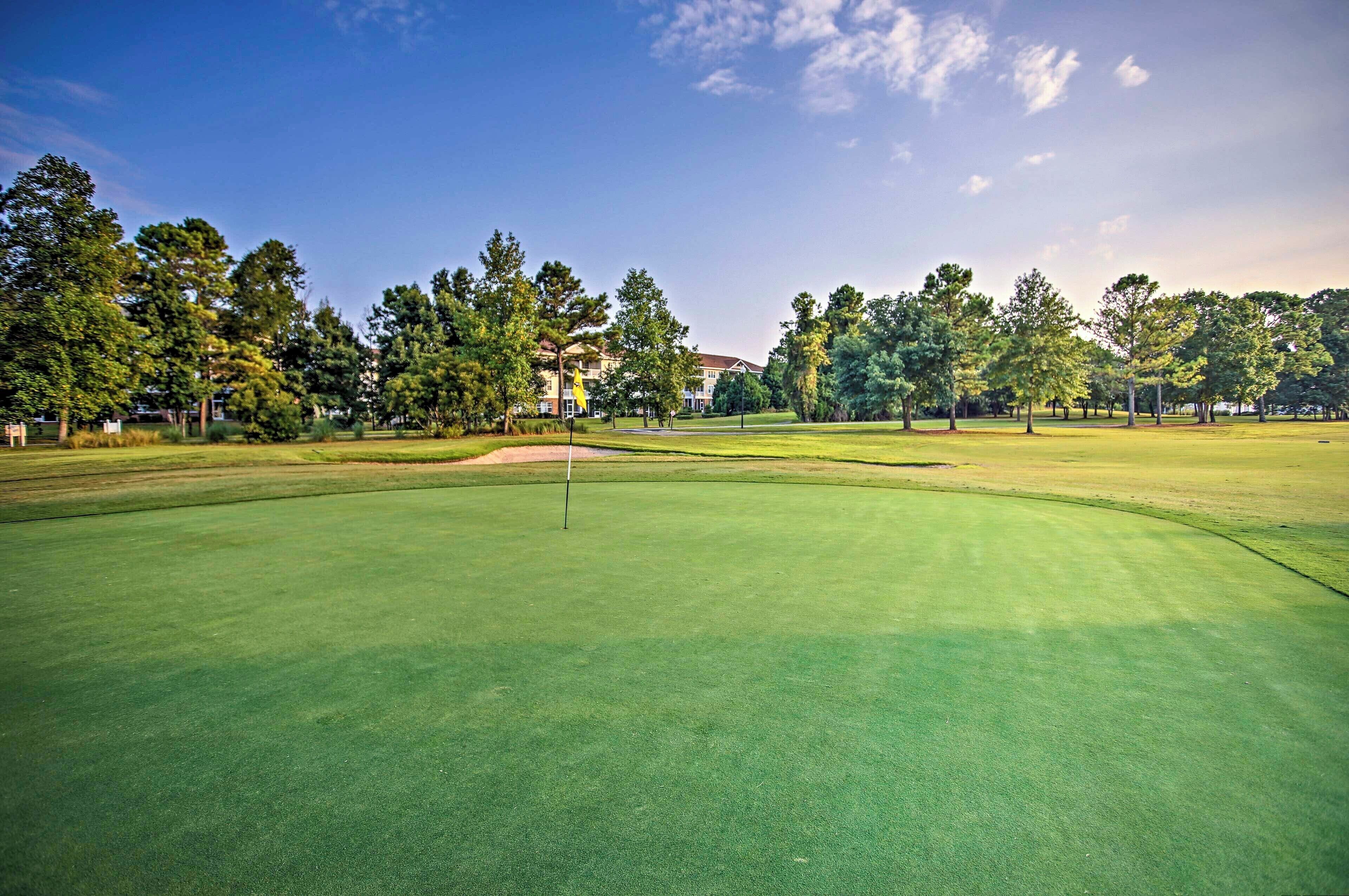 A golf course with a flagstick, surrounded by trees and a building in the background.