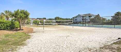 A sandy volleyball court with a net, surrounded by greenery and a building in the background.