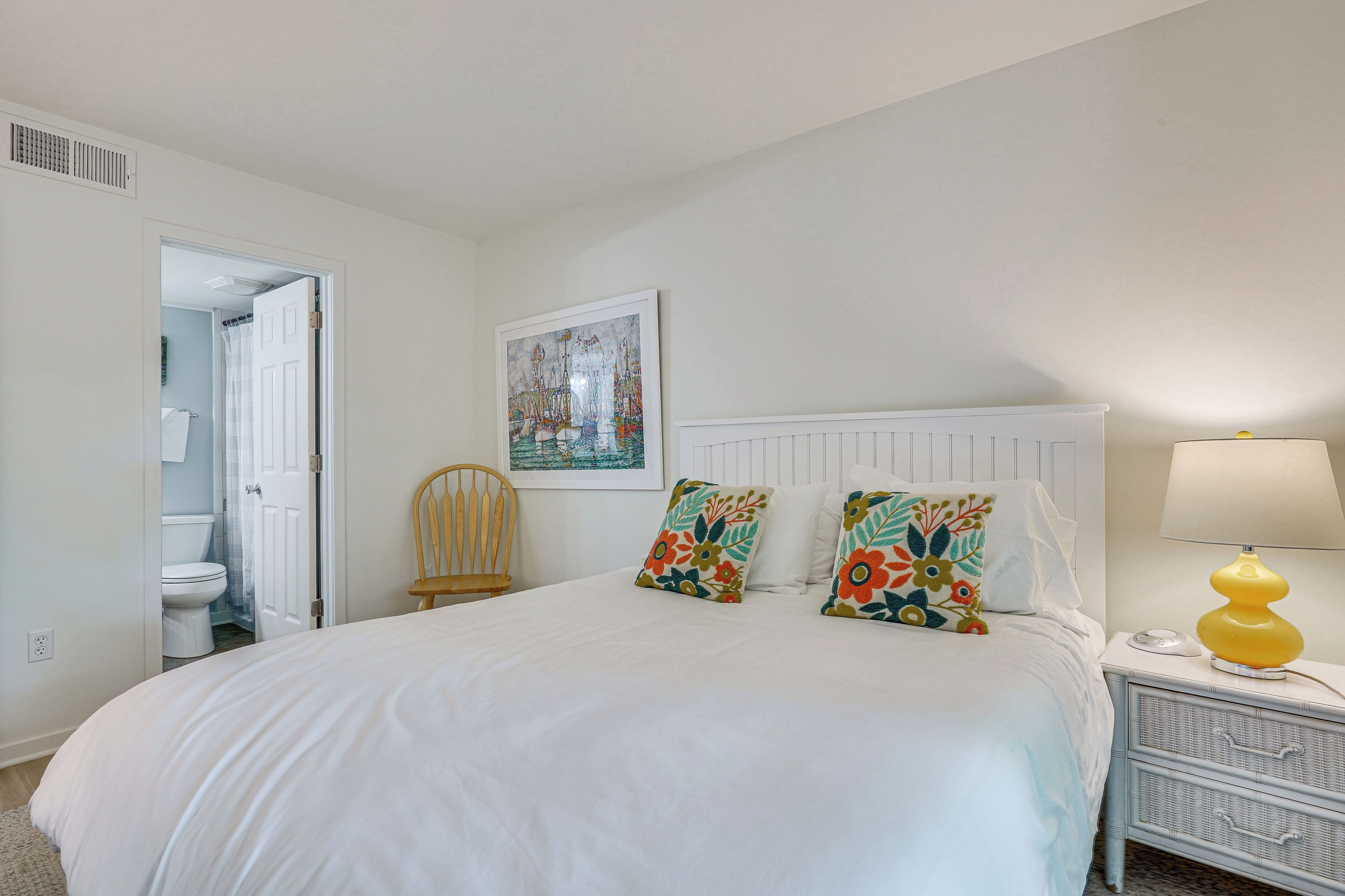 A bedroom with a bed, a nightstand with a lamp, a chair, and a painting on the wall.