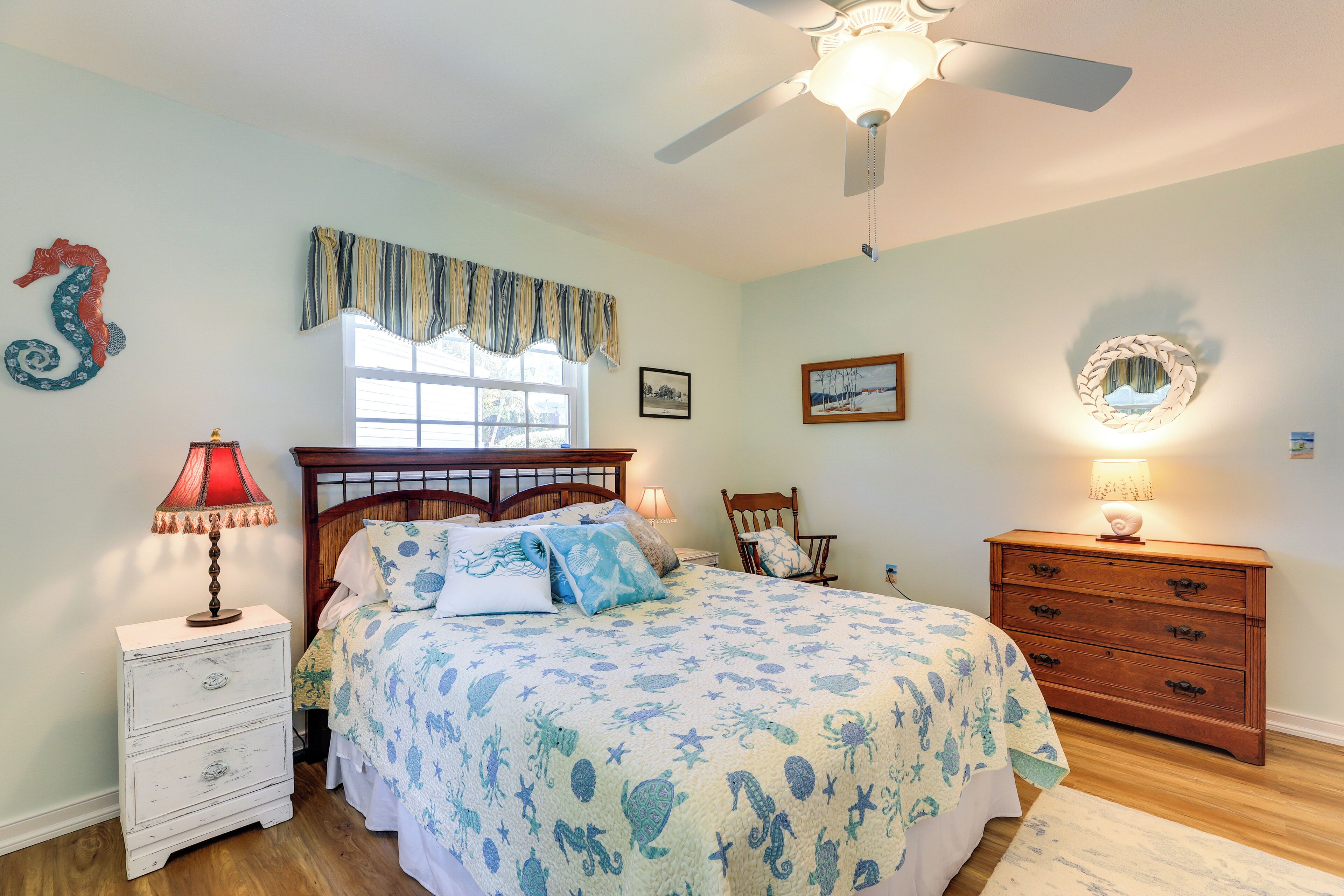 Un dormitorio con cama, mesita de noche, cómoda, ventilador de techo y ventana con cortinas.