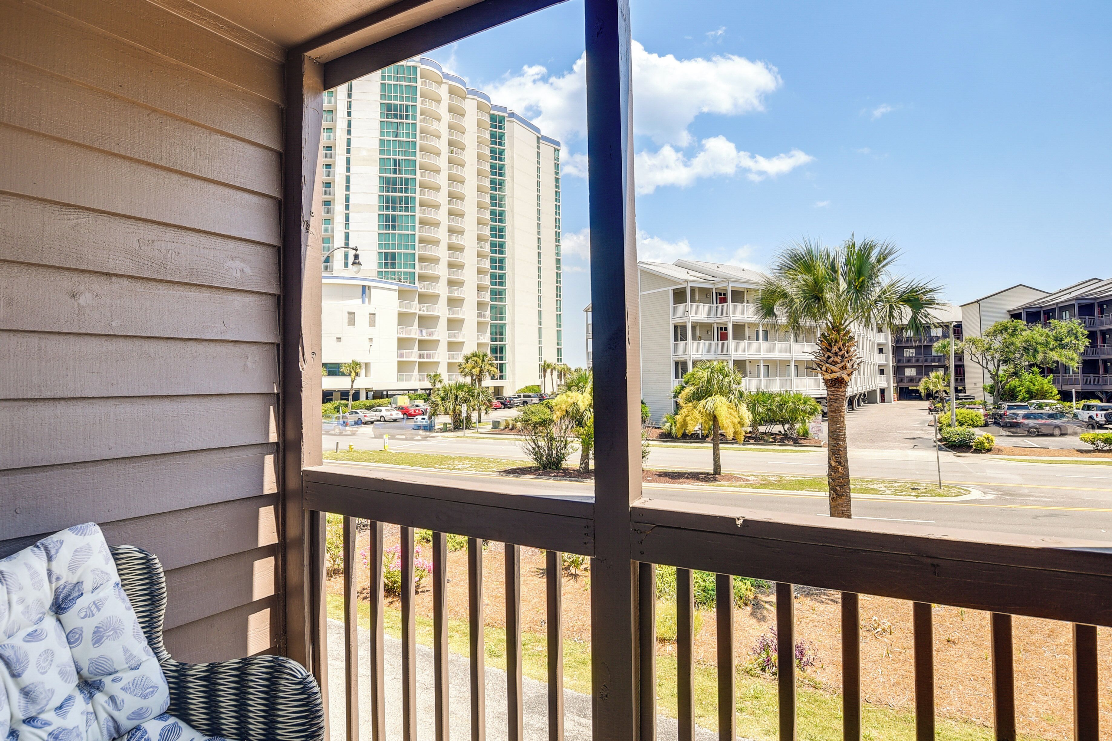 A balcony with a view of a multi-story building, palm trees, and a parking lot.
