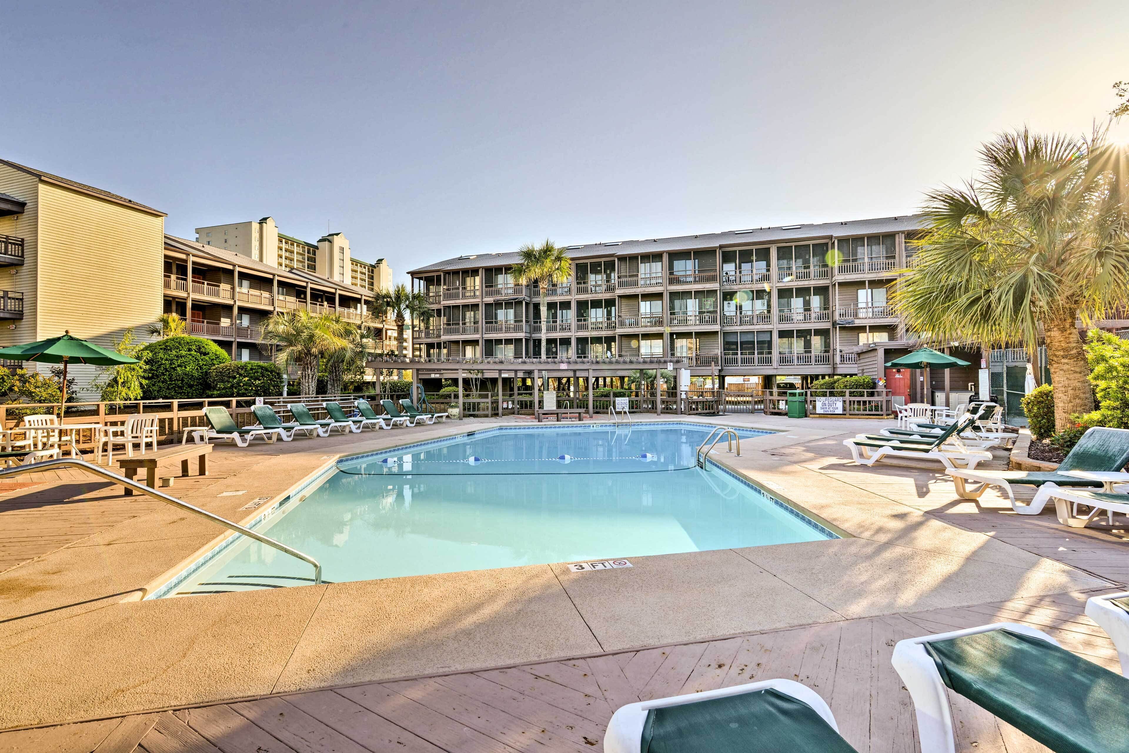 A hotel pool area with lounge chairs, a swimming pool, and a multi-story building with balconies.