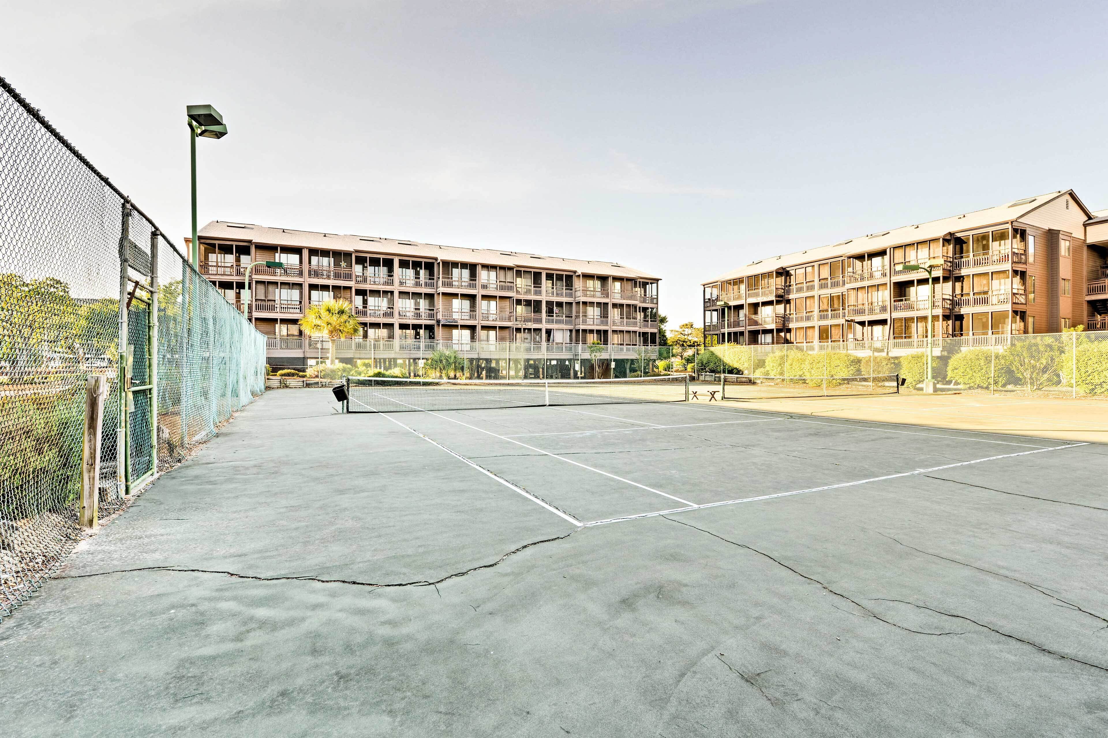 An outdoor tennis court with a net, surrounded by a chain-link fence, and adjacent multi-story buildings.