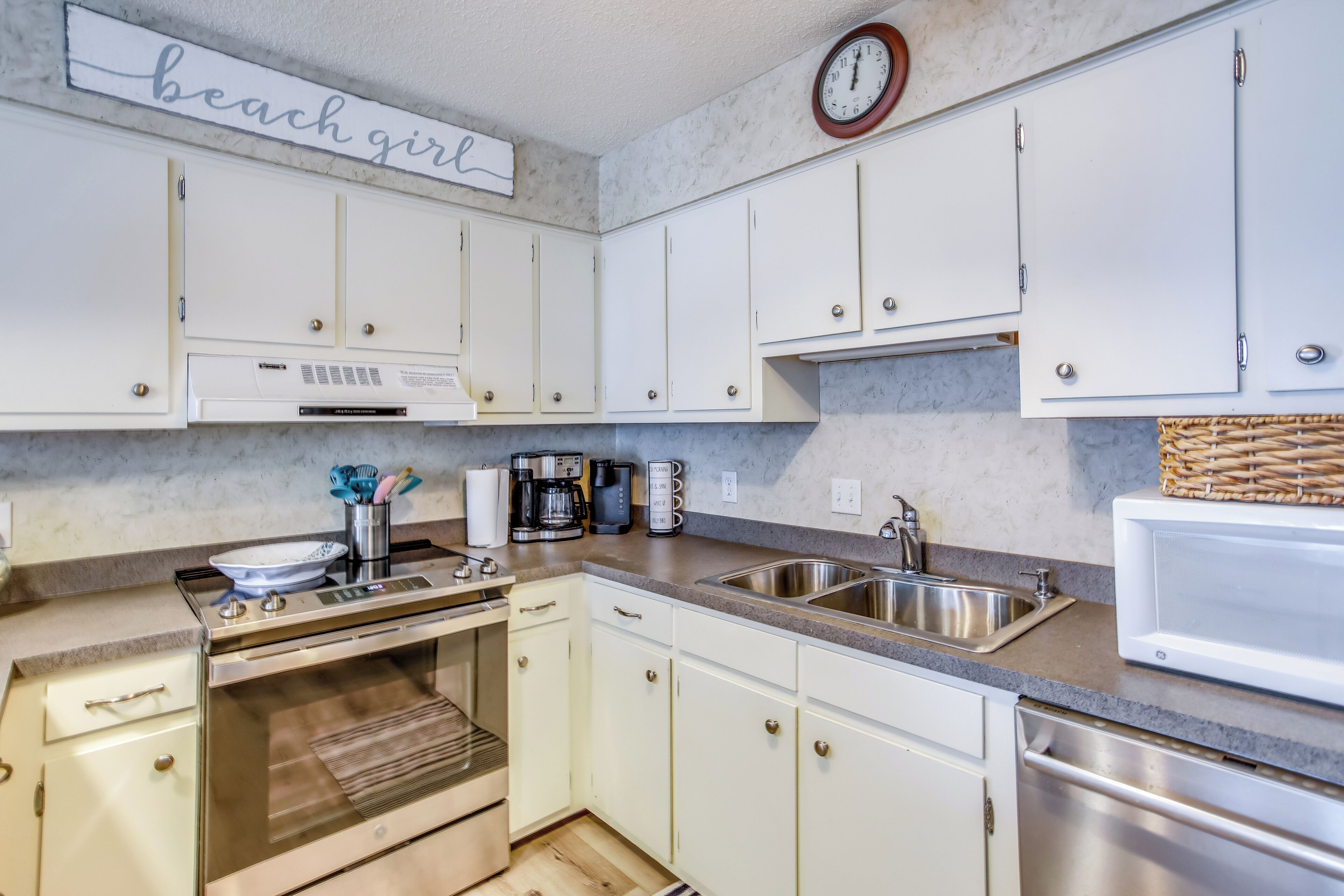 A kitchen with white cabinets, stainless steel appliances, a clock, and a 'beach girl' sign.