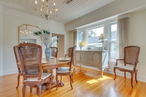 A dining room with a wooden table, four chairs, a chandelier, and a large window with curtains. - Pensacola Paradise: Furnished Deck & Gas Grill! (Pensacola)