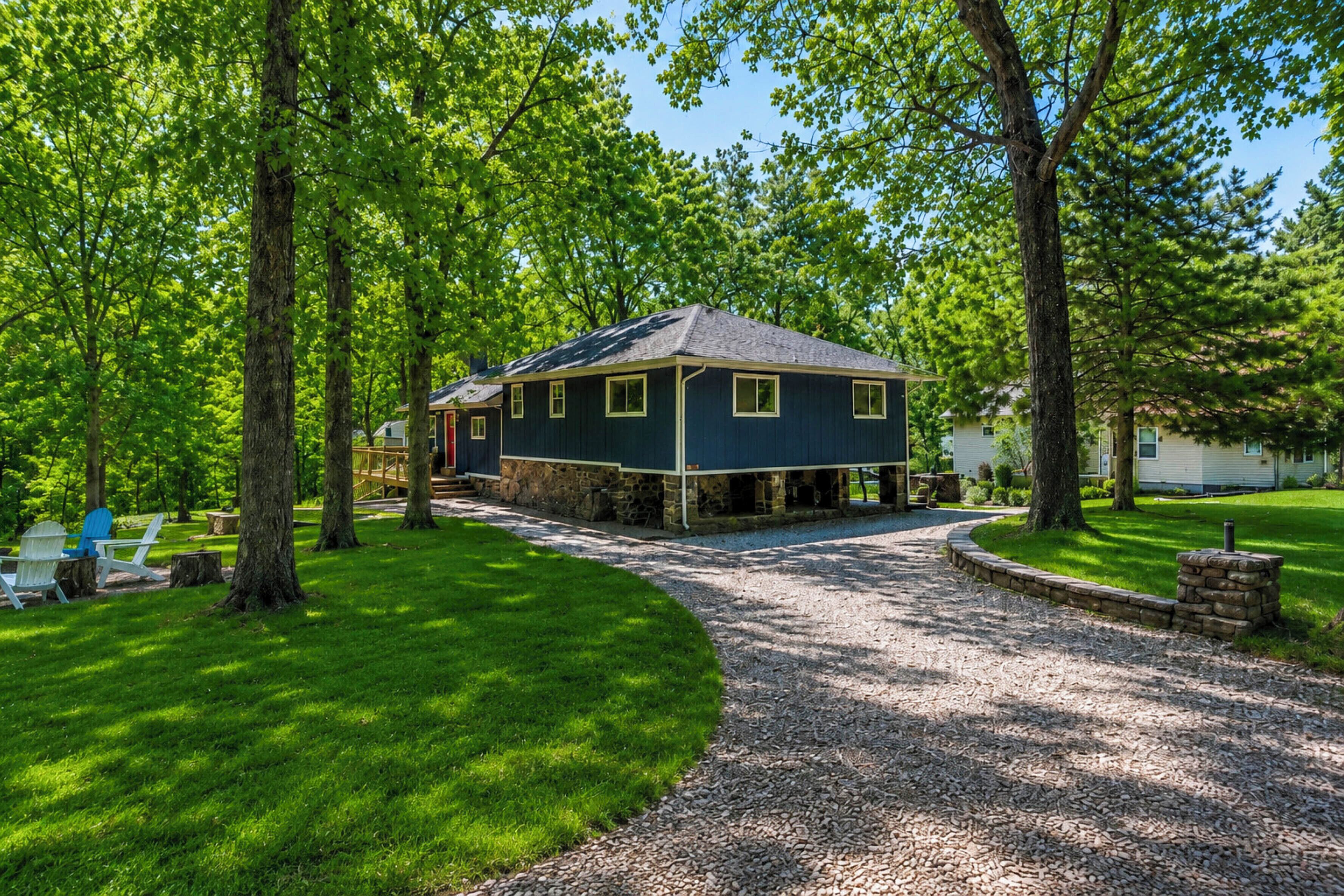 Une maison avec des fondations en pierre, une façade d’un bleu foncé et une allée en gravier menant à une terrasse en bois.