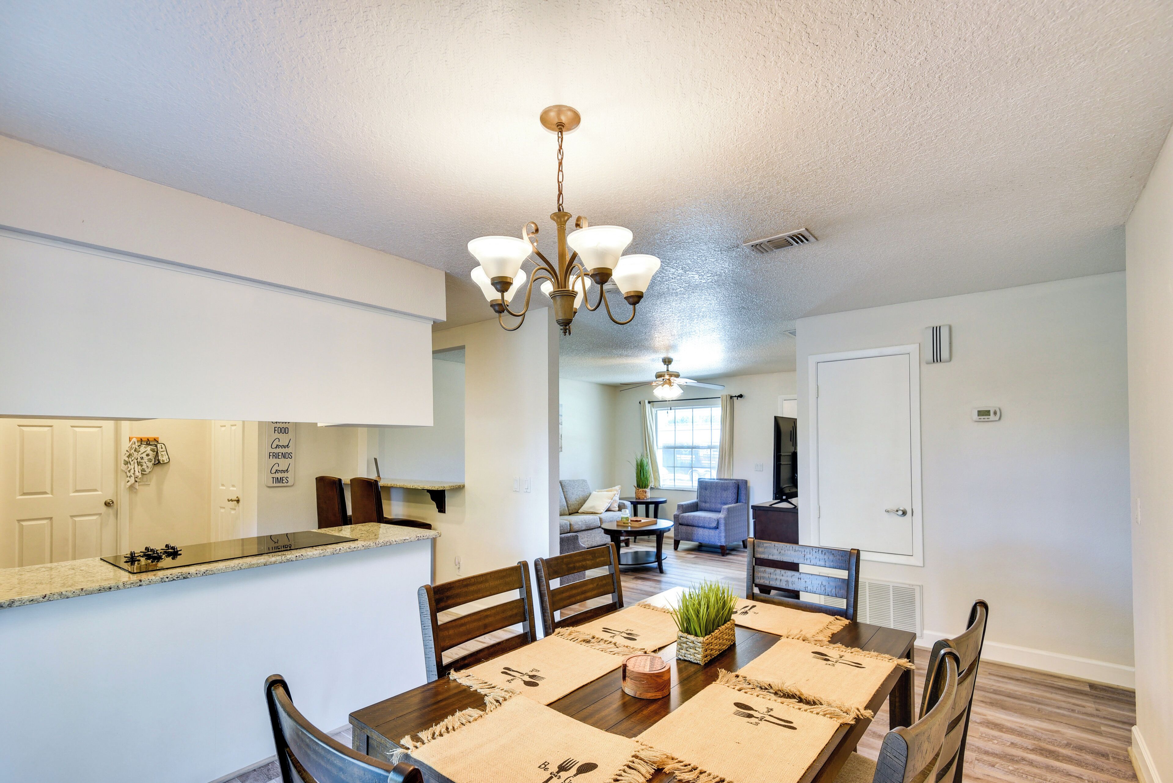 Uma área de refeições com mesa de madeira, cadeiras e um candelabro. É visível uma área de cozinha com bancada e armários ao fundo.