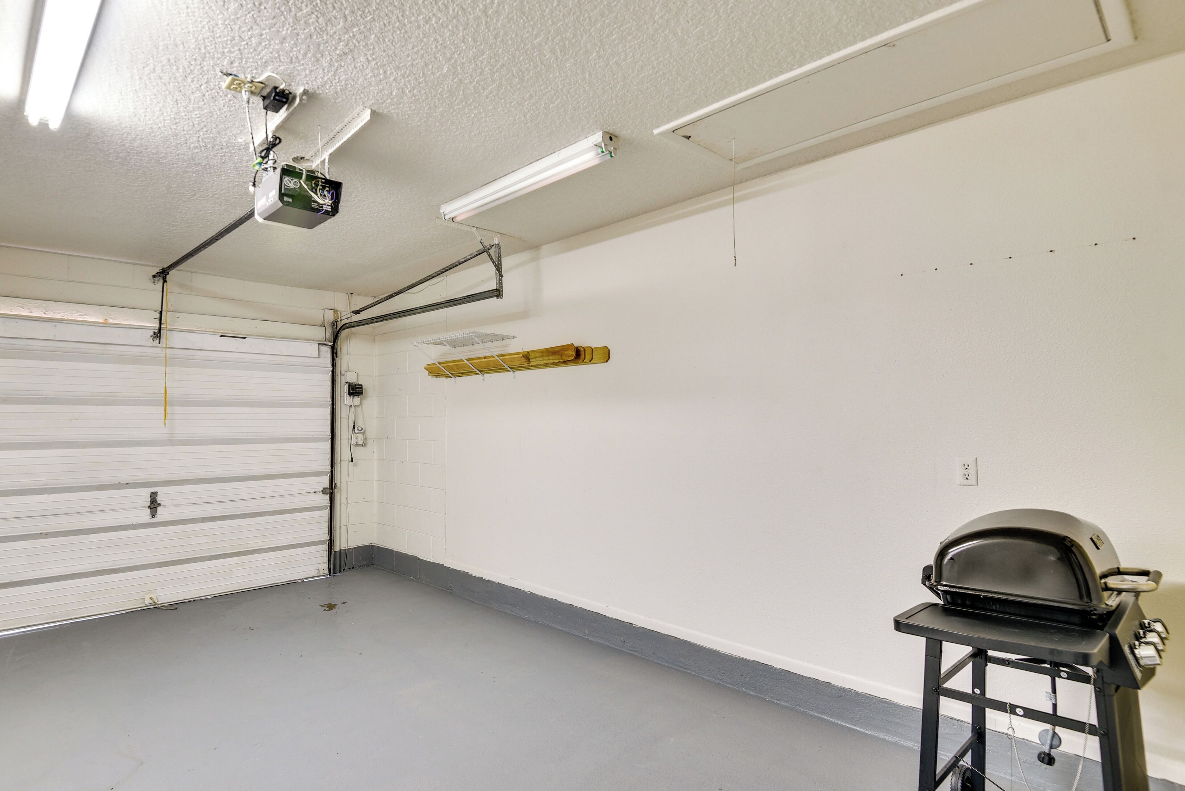 A garage with a white garage door, fluorescent lighting, a ceiling-mounted projector, and a barbecue grill on a cart.