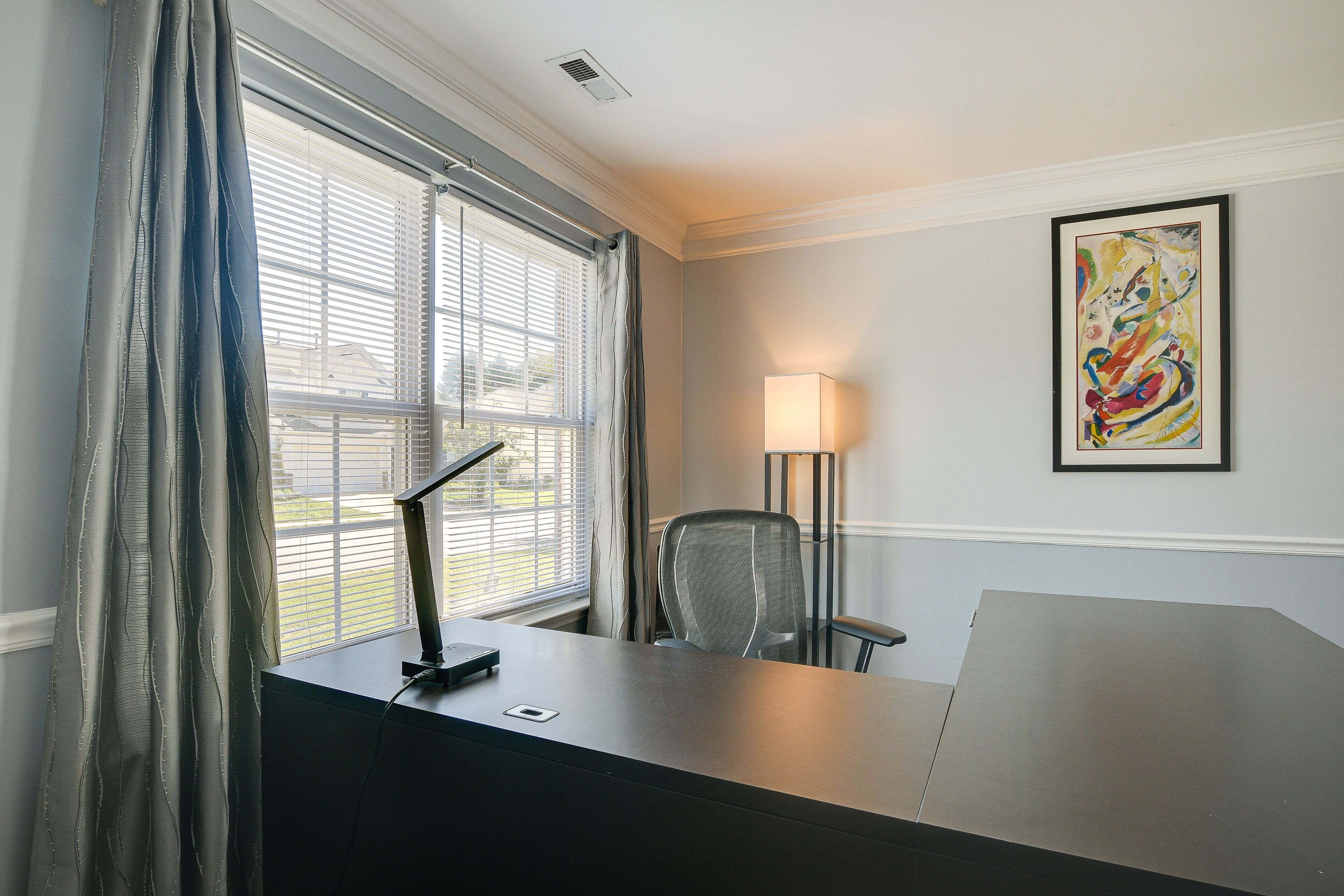Habitación de hotel con escritorio, silla, ventana con persianas, lámpara y un cuadro abstracto enmarcado en la pared.