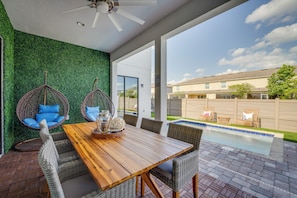 Um pátio com mesa de madeira e cadeiras de vime, uma cadeira suspensa com almofadas azuis, e vista para uma piscina e edifícios ao fundo.