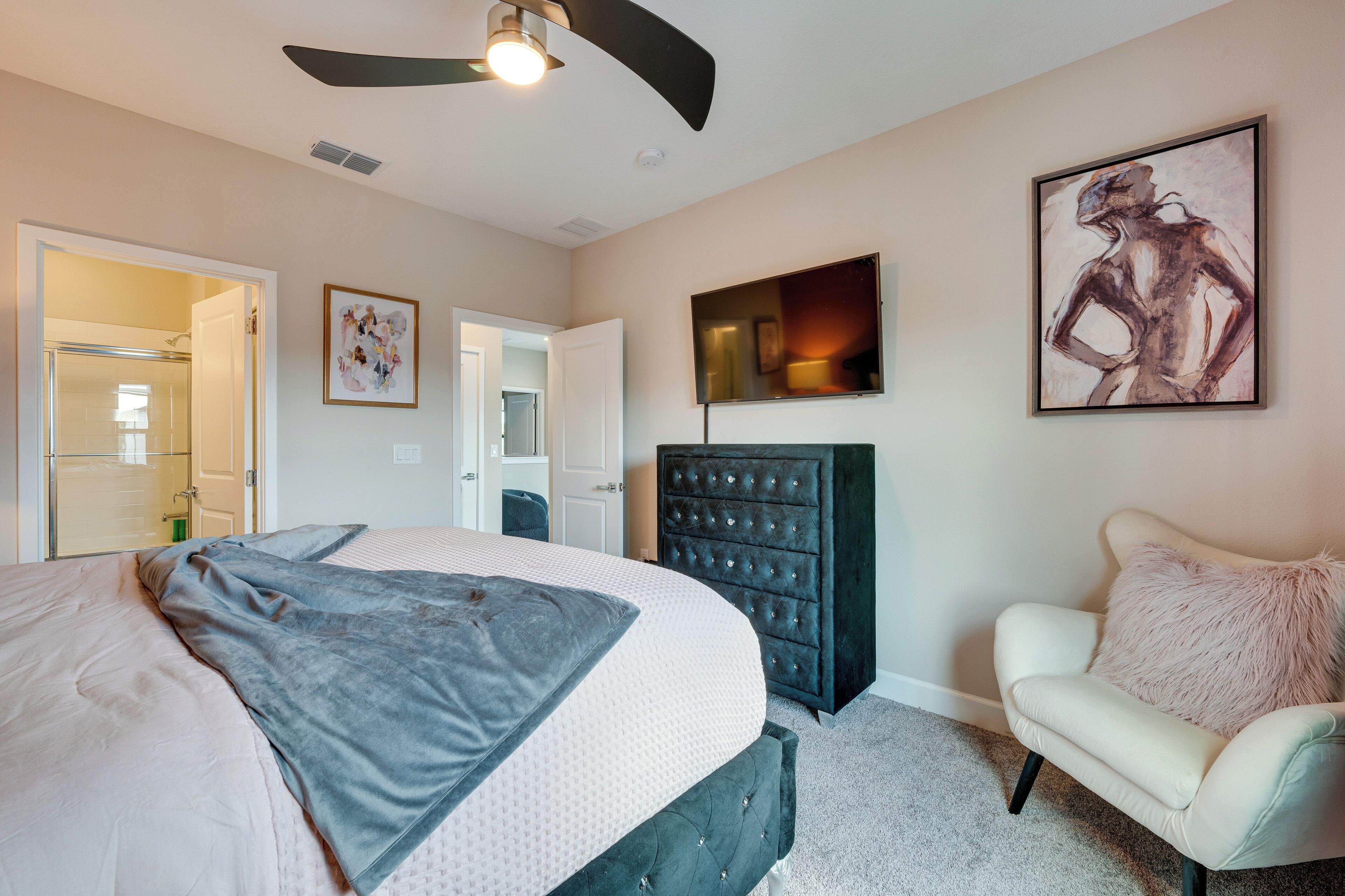 Une chambre à coucher avec un lit, une commode, une chaise, un ventilateur de plafond et un tableau accroché au mur.