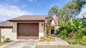 A residential house with a brown garage door, a concrete driveway, and a well-maintained garden with various plants and an American flag. - Walk to Restaurants: Downtown Leesburg Townhome! (Leesburg)