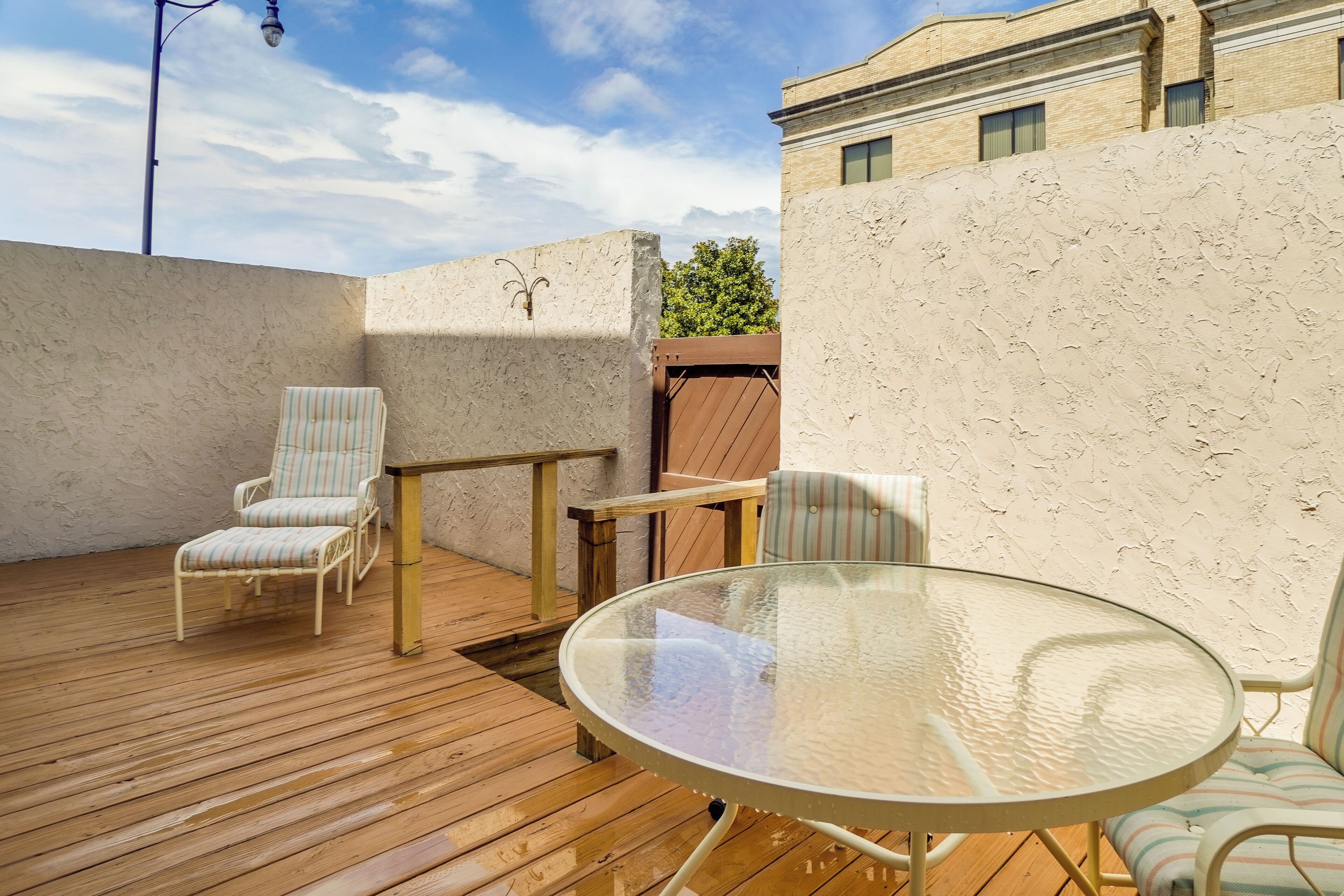 Une terrasse extérieure avec une table en verre et des chaises à rayures, entourée de murs à motifs et d’une plateforme en bois.