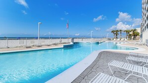 A swimming pool with lounge chairs and an American flag, surrounded by a fence and palm trees, with a clear view of the ocean. - Beachfront Condo in Gulf Shores w/ Pool Access! (Gulf Shores)