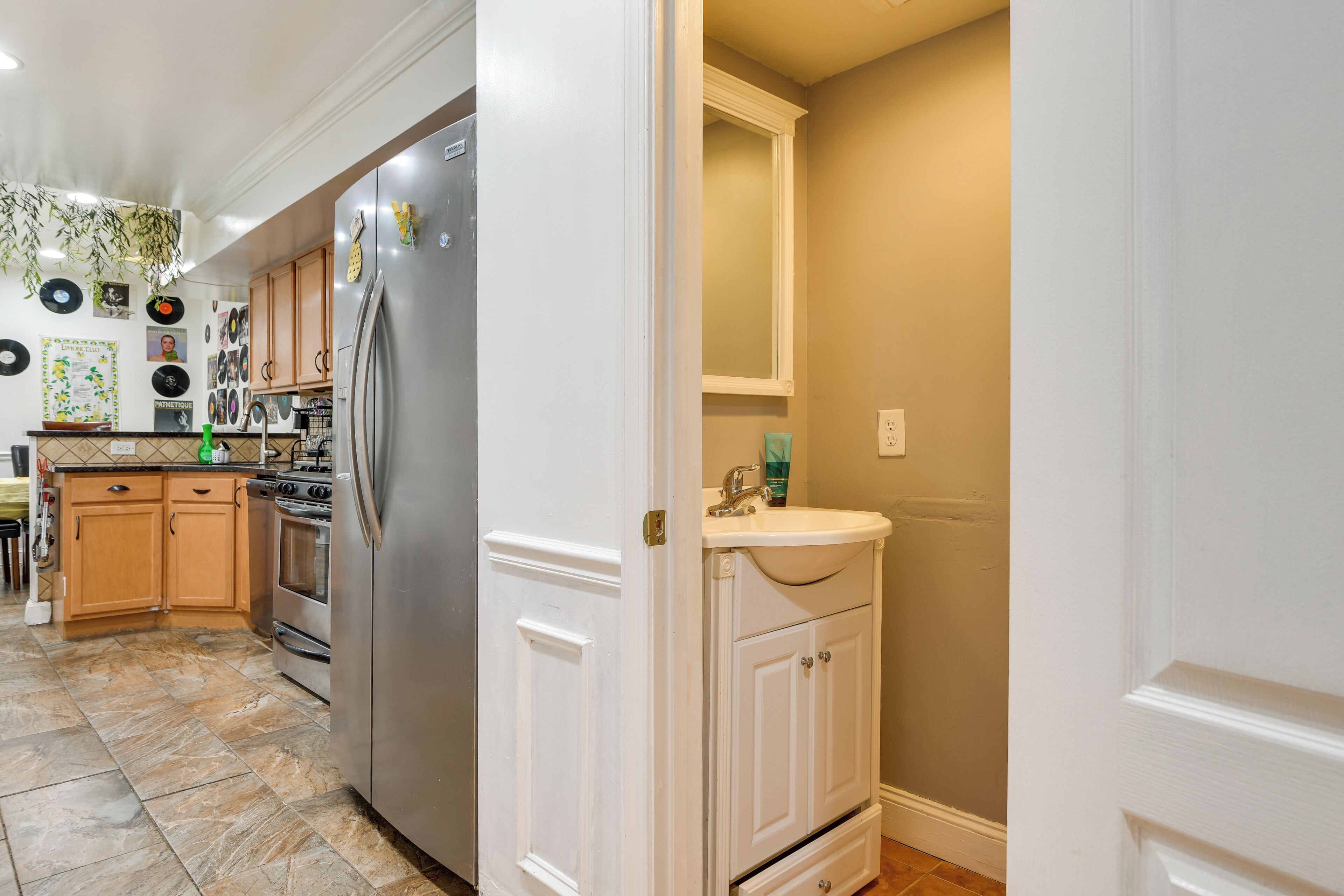 A kitchen with a stainless steel refrigerator, wooden cabinets, a tiled floor, and a white door leading to a bathroom.