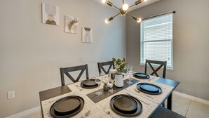 A dining area with a table set for four, featuring dark plates, glasses, and a vase with flowers. There are wall-mounted artworks and a ceiling light fixture.