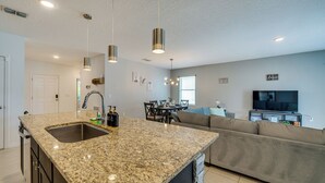 A modern kitchen with granite countertops, a double sink, and pendant lighting. There is a dining area with a table and chairs, and a living area with a sofa and a flat-screen TV.