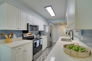 A modern kitchen with white cabinets, stainless steel appliances, a microwave, oven, and a refrigerator. There is a clock on the wall and a basket of green apples on the counter.