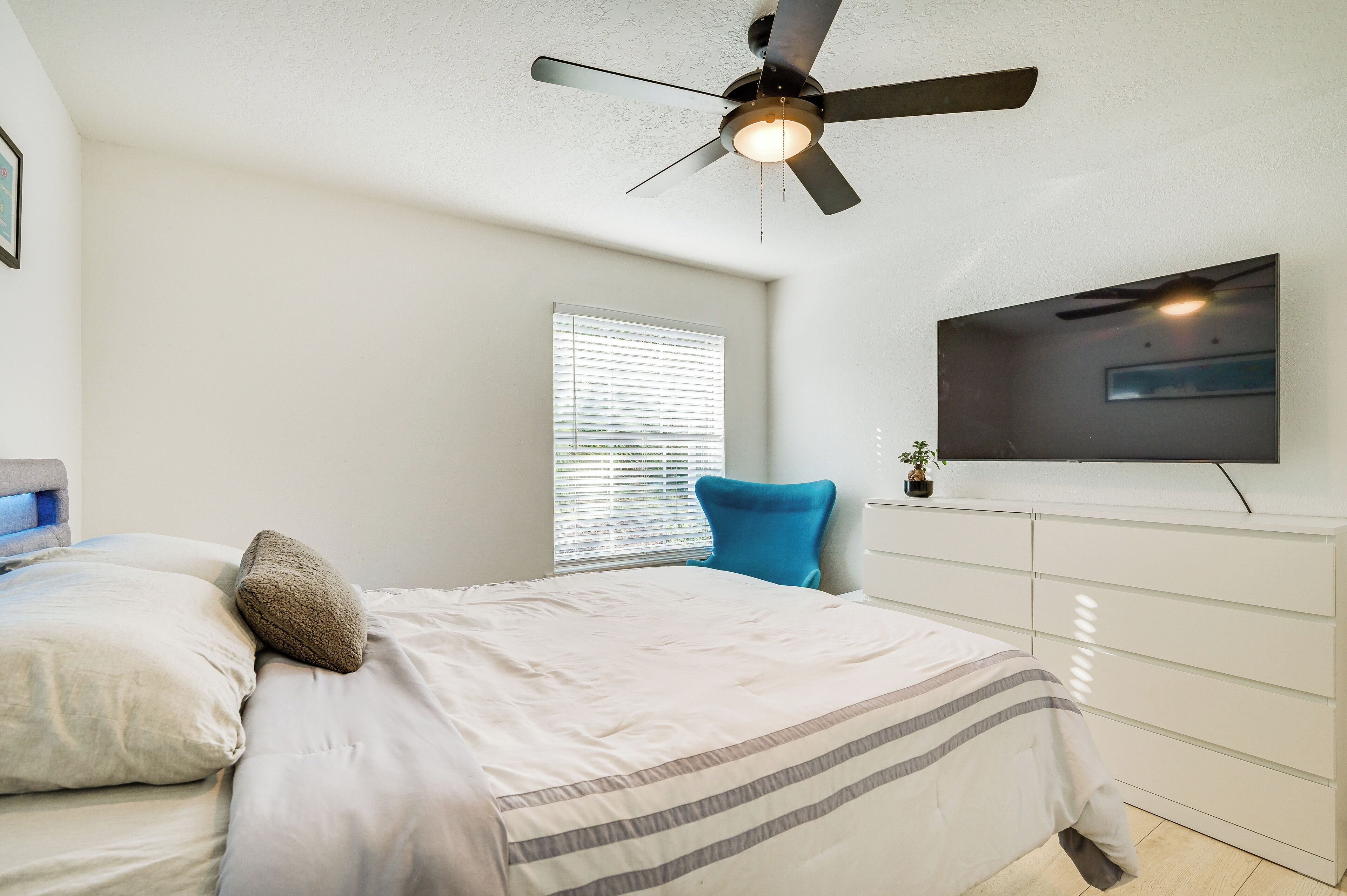 A bedroom with a bed, a ceiling fan, a window with blinds, a TV mounted on the wall, and a dresser.