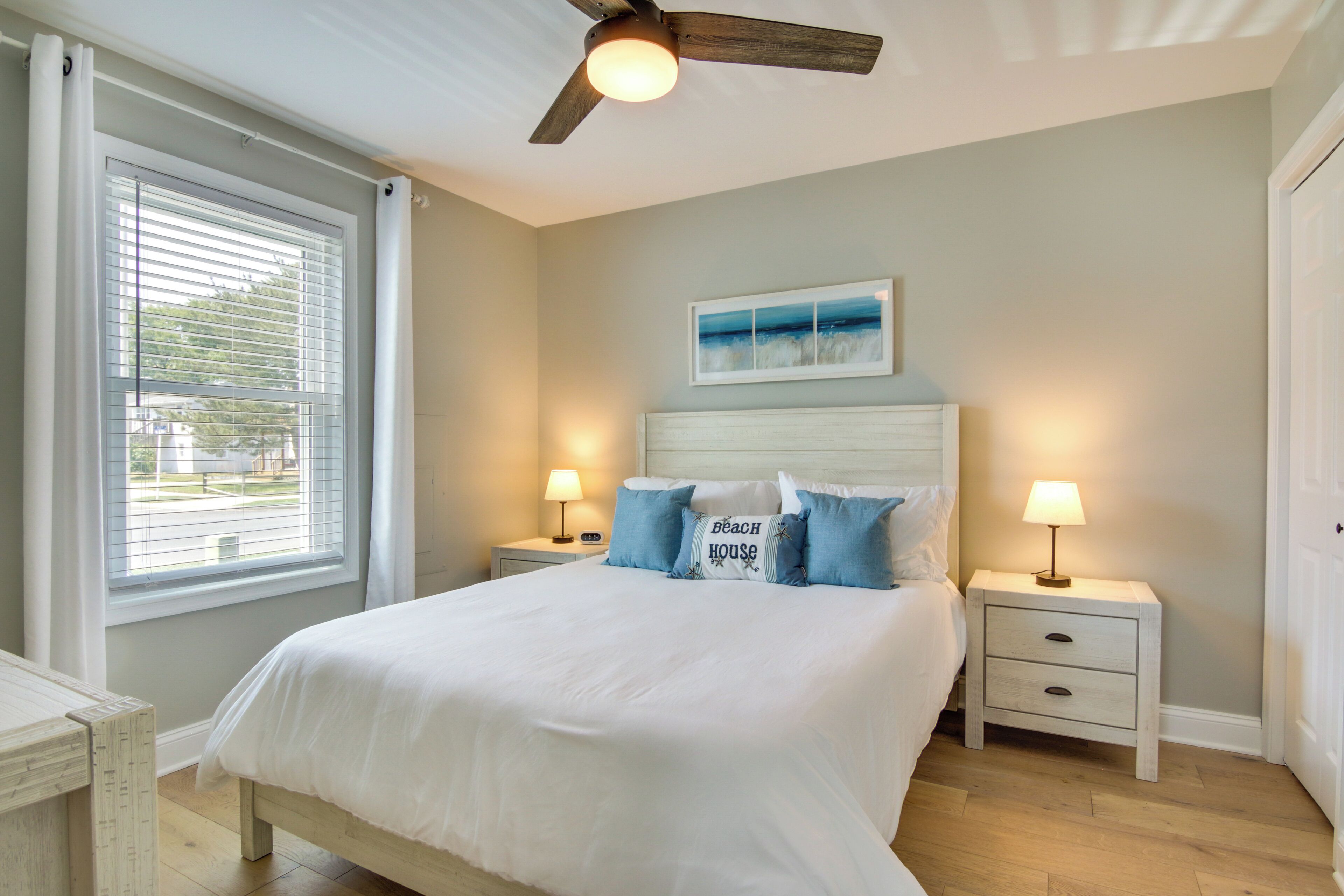 A bedroom with a bed, two bedside tables with lamps, a ceiling fan, a window with blinds, and a painting on the wall.