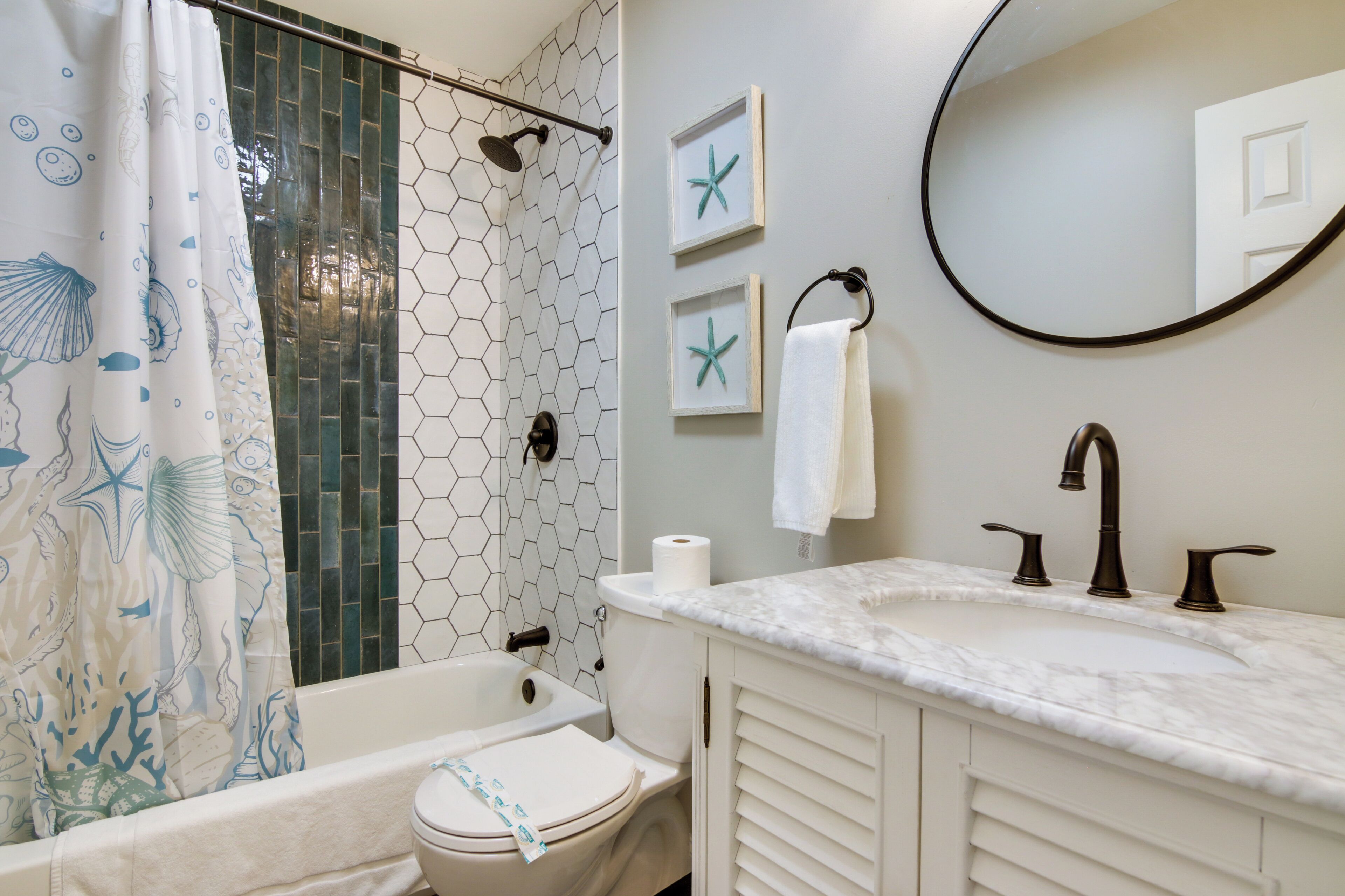 Une salle de bain avec un mur recouvert de carreaux hexagonaux, une baignoire, des toilettes, un lavabo doté d’un plan de travail en marbre et des décorations murales représentant des étoiles de mer encadrées.