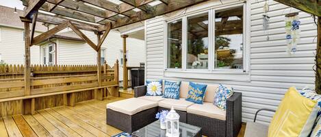 A covered patio with a wooden deck, a wicker sofa with cushions, and a glass coffee table.