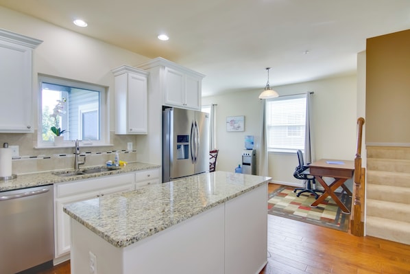 Cocina moderna con armarios blancos, encimeras de granito, un refrigerador de acero inoxidable y un comedor con mesa y sillas de madera.