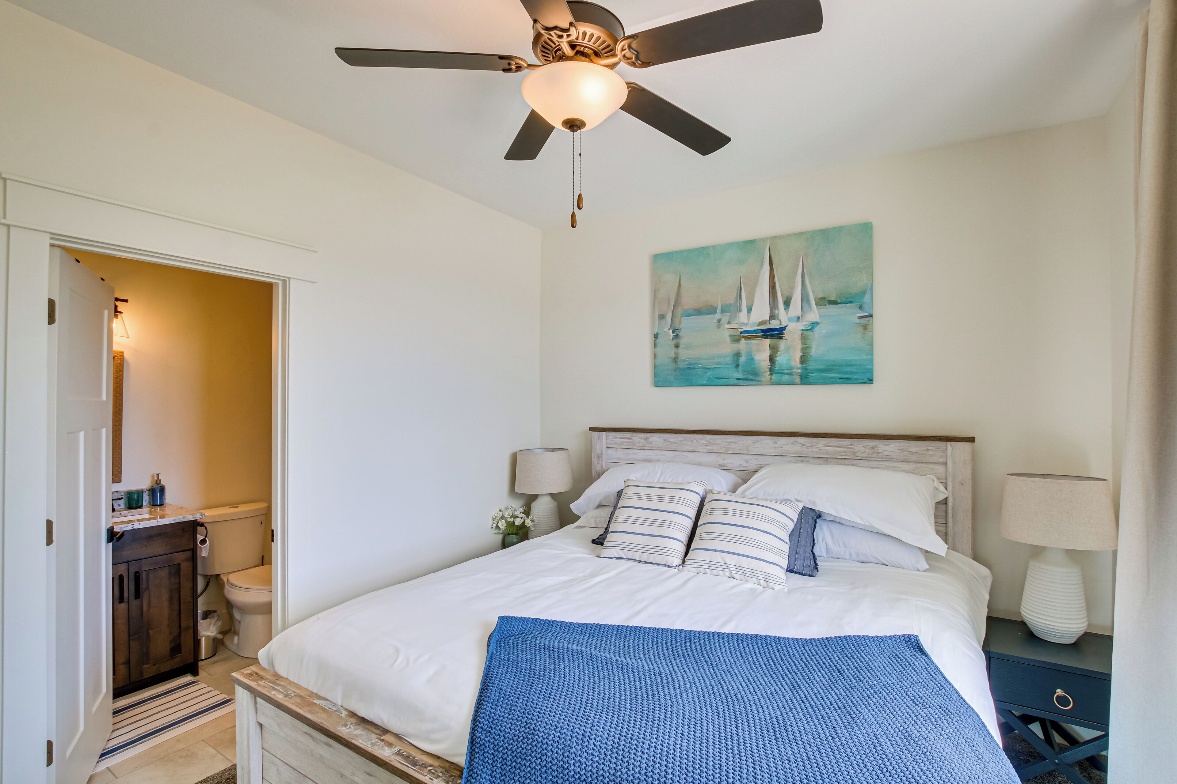 Quarto com cama, ventilador de teto, pintura de veleiros, mesa de cabeceira com abajur e um banco de madeira.