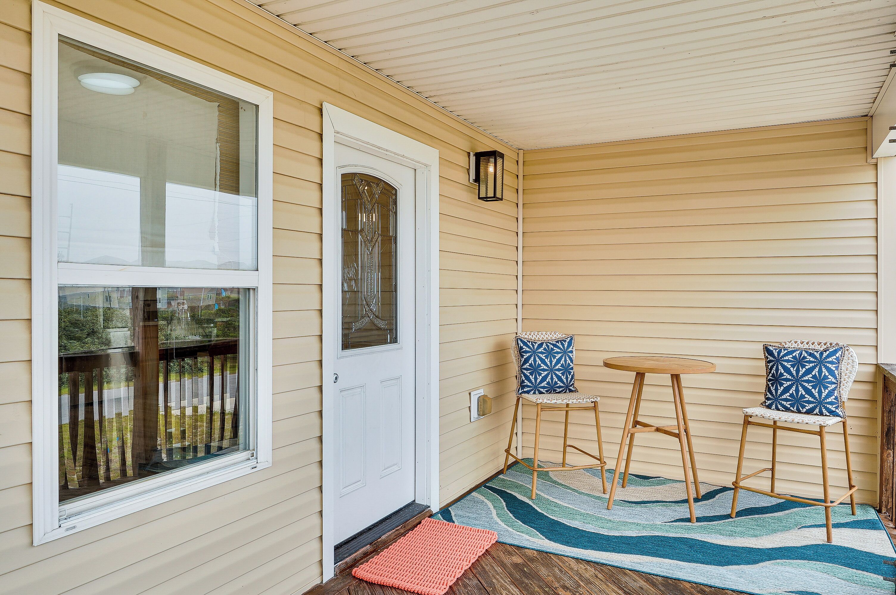 A porch with a white door, two chairs with patterned cushions, a small round table, and a colorful rug.