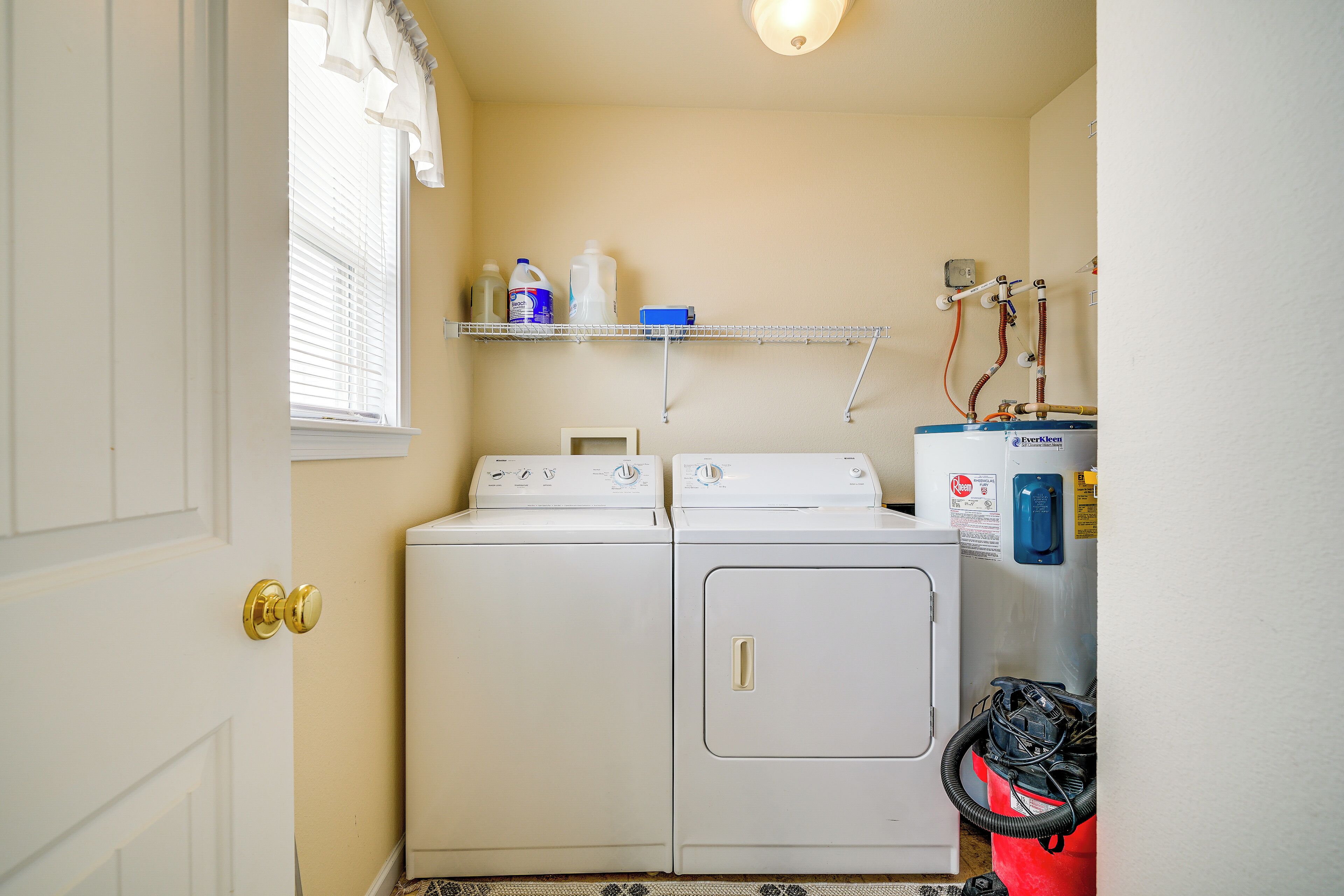 A laundry room with a front-loading washer and dryer, a shelf with cleaning supplies, and a water heater with hoses.