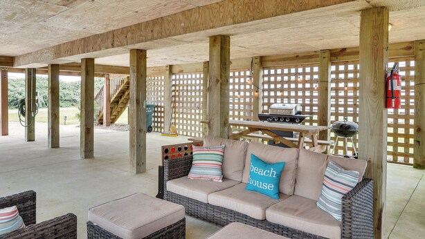 A covered patio area with a wicker sofa, cushions, and a wooden table with a grill. - Nags Head Vacation Rental w/ Hot Tub: Near Beach! (Nags Head)