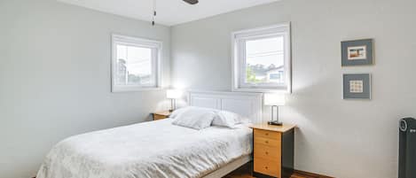 Un dormitorio con una cama, dos mesitas de noche, un ventilador de techo y dos ventanas.