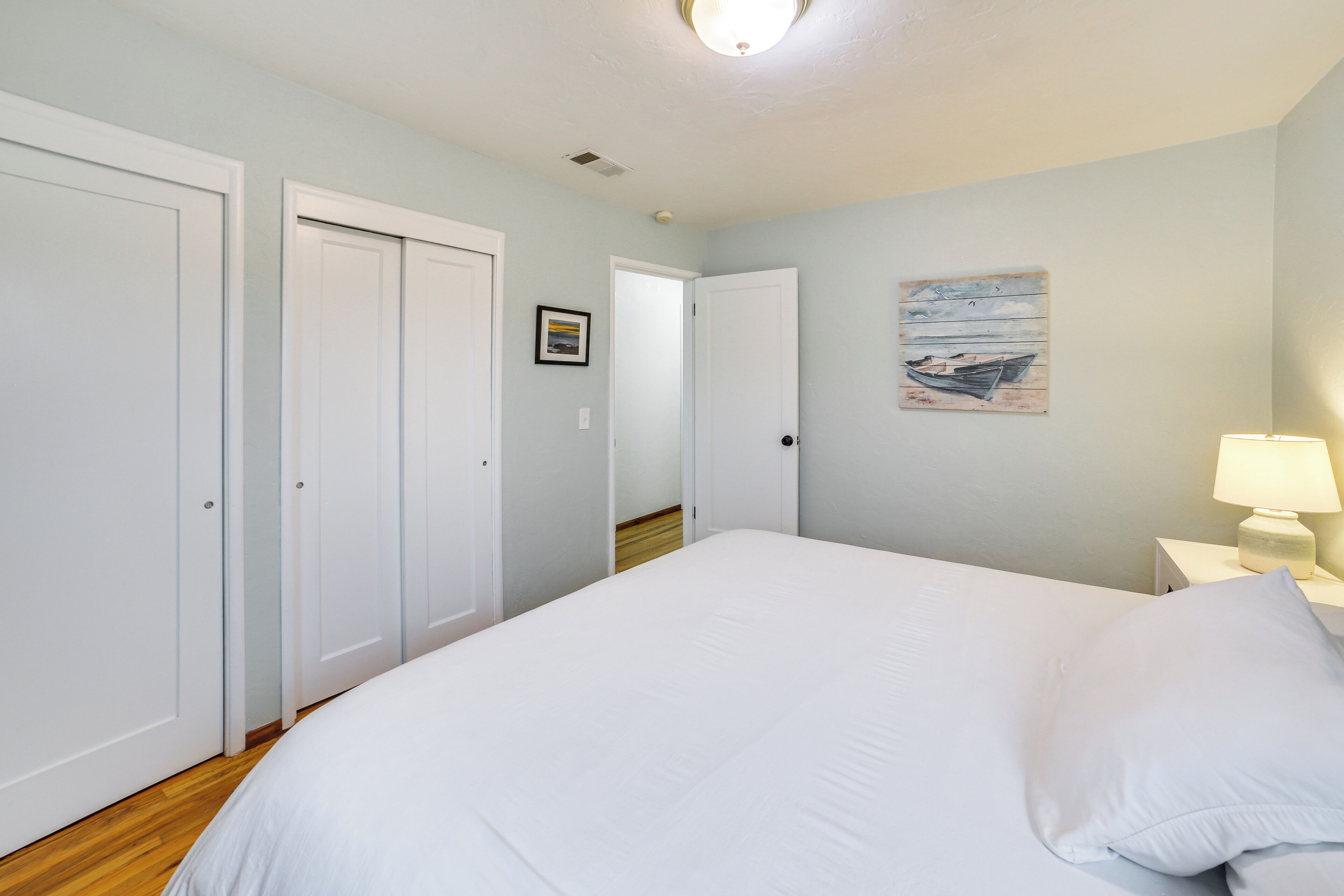 A bedroom with a large bed, white walls, and a wooden floor. There are white doors and a nightstand with a lamp.