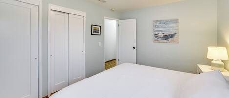 A bedroom with a large bed, white walls, and a wooden floor. There are white doors and a nightstand with a lamp.