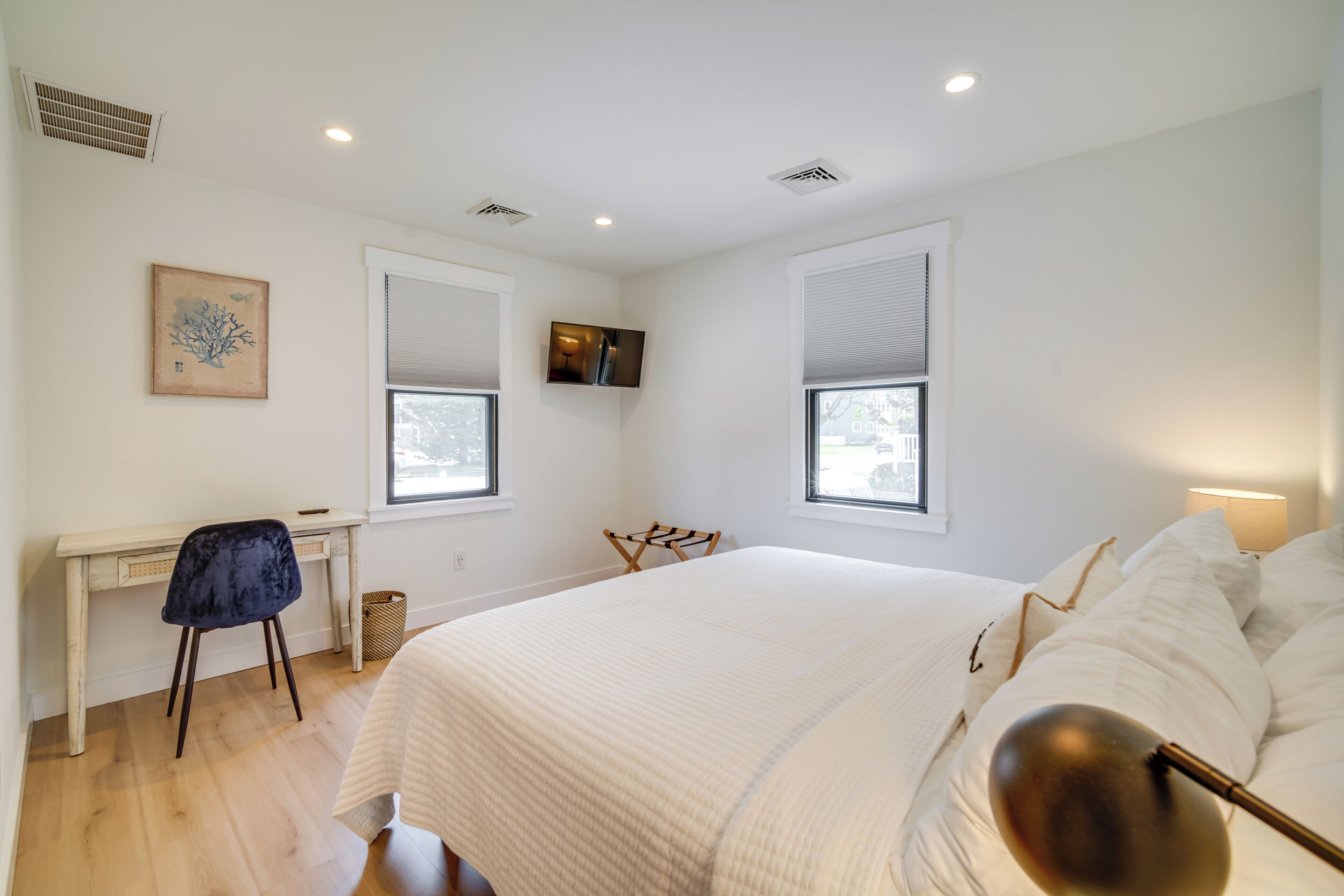 A bedroom with a large bed, a desk, and a chair. There are two windows and a TV mounted on the wall.