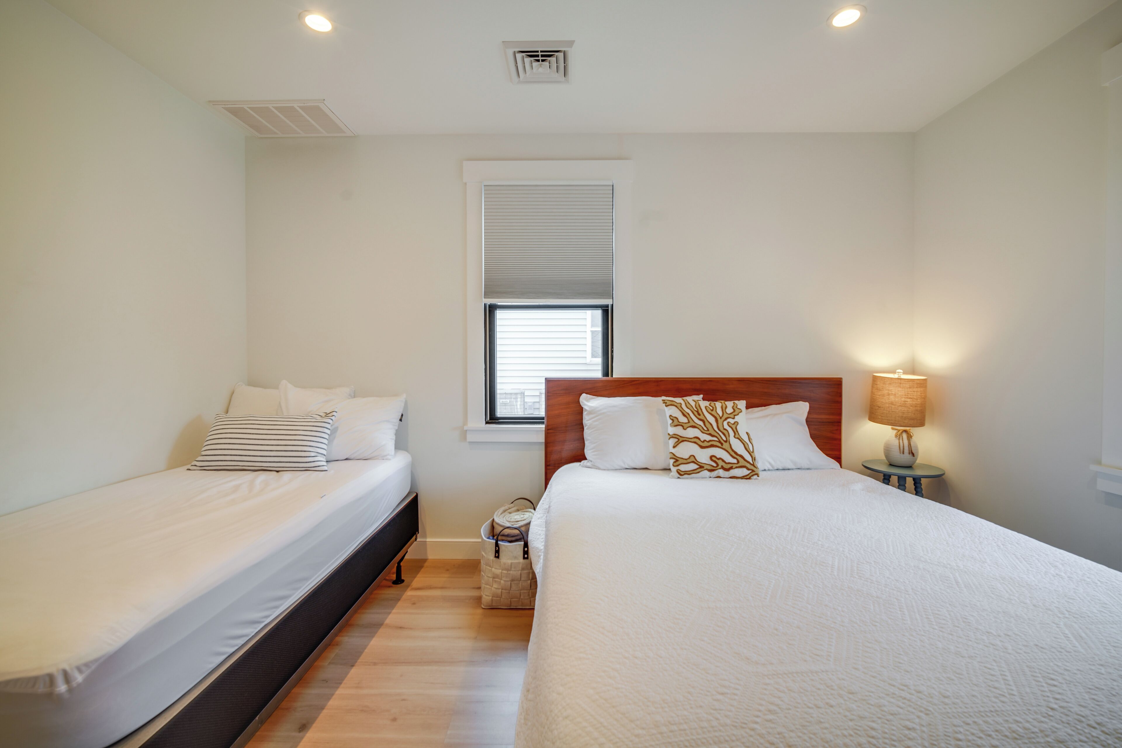 A bedroom with two single beds, a window with blinds, a nightstand with a lamp, and a ceiling vent.