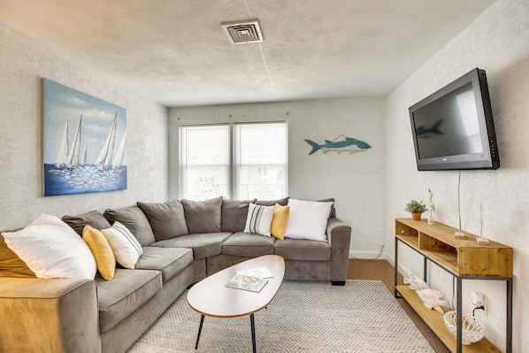 A living room with a gray sectional sofa, a small wooden coffee table, a TV mounted on the wall, and a painting of sailboats.