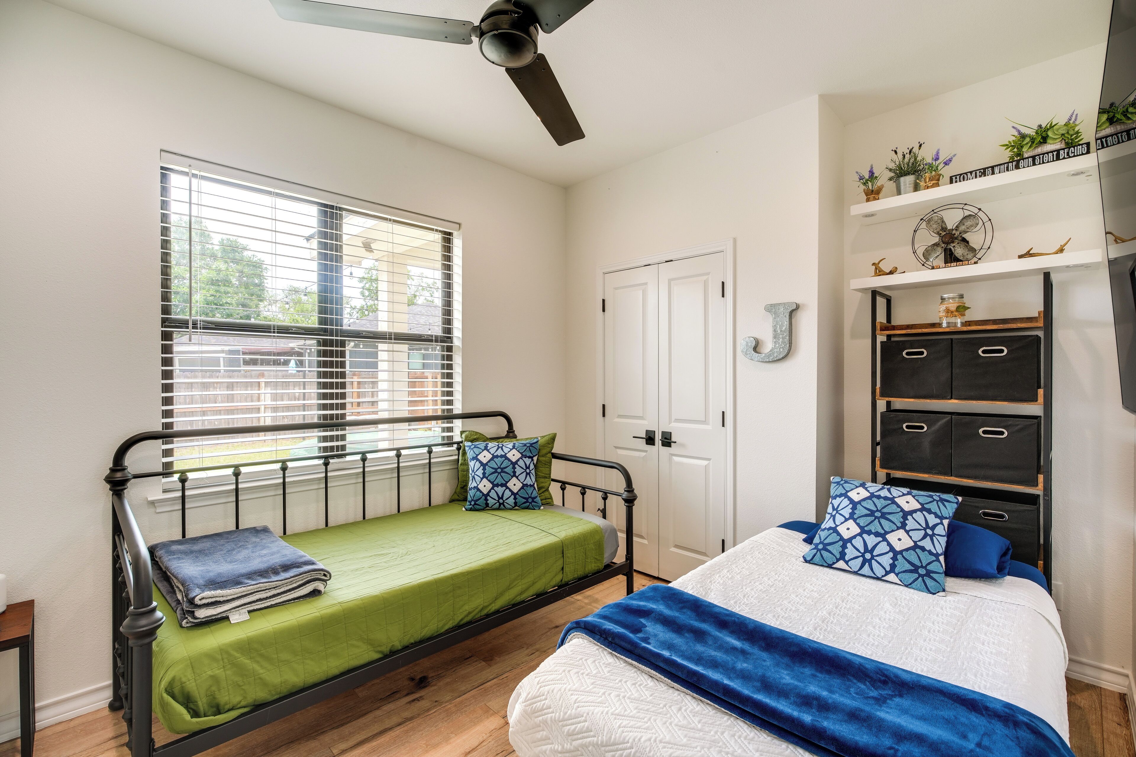 Quarto com uma cama de solteiro, janela com persianas, ventilador de teto, guarda-roupa e prateleira com objetos decorativos.