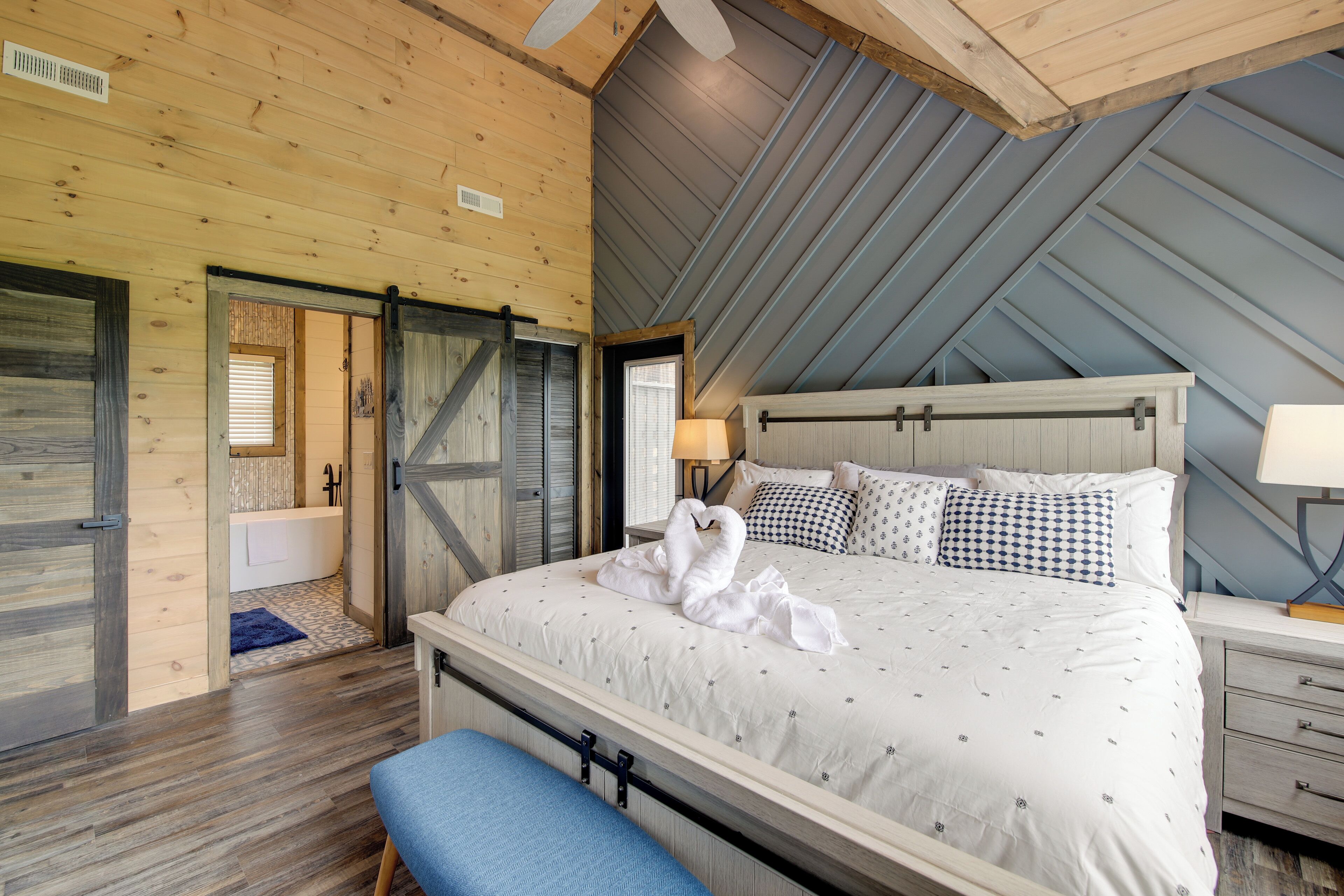 A bedroom with a bed, a bench, a nightstand, a lamp, and a view of the bathroom.