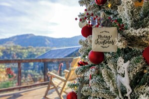 Een kerstboom met versieringen en een bord met de tekst 'Merry Christmas', een houten stoel en een uitzicht op bergen.