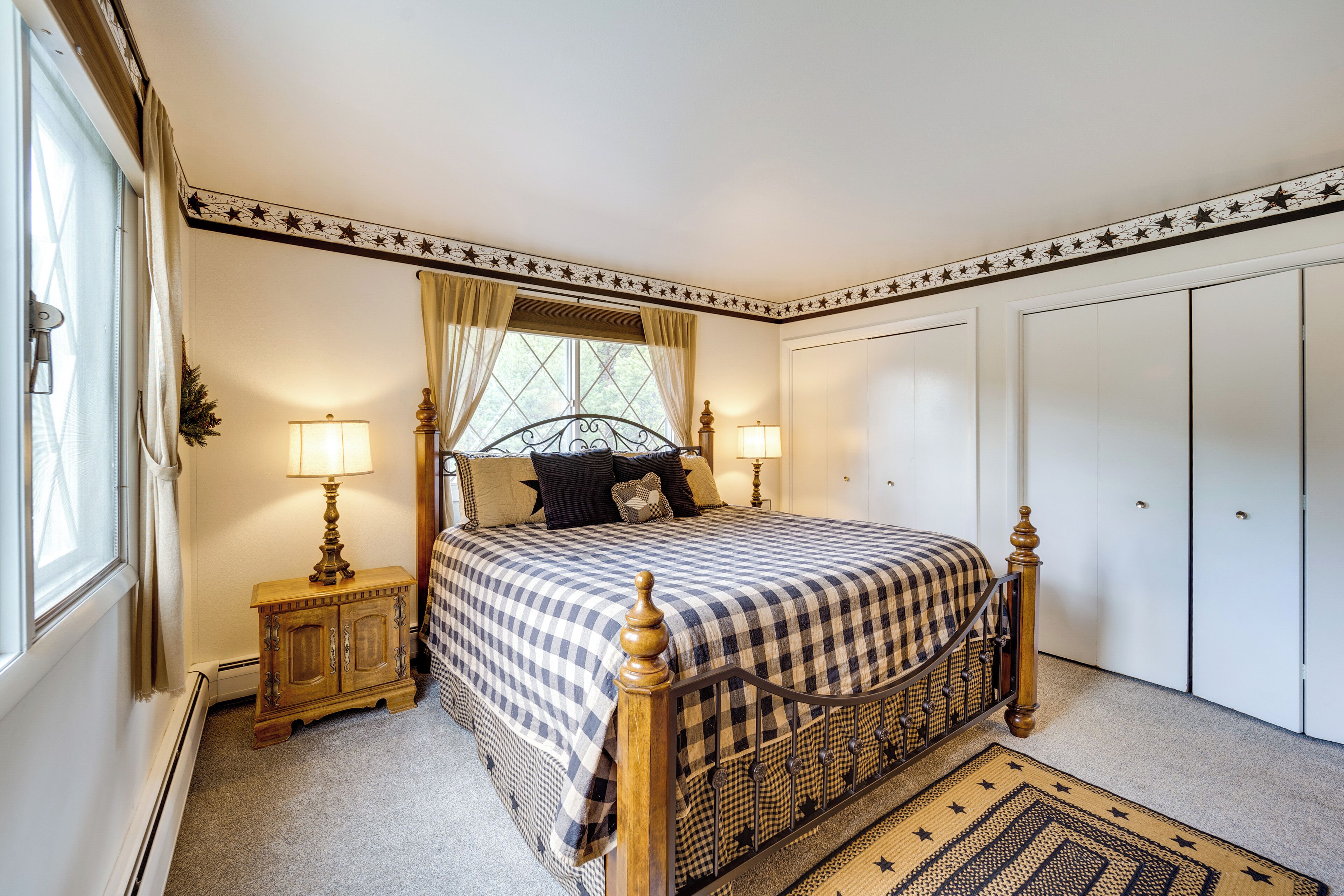 A bedroom with a checkered bedspread, two bedside lamps, a small wooden cabinet, a star-patterned ceiling border, and a window with curtains.