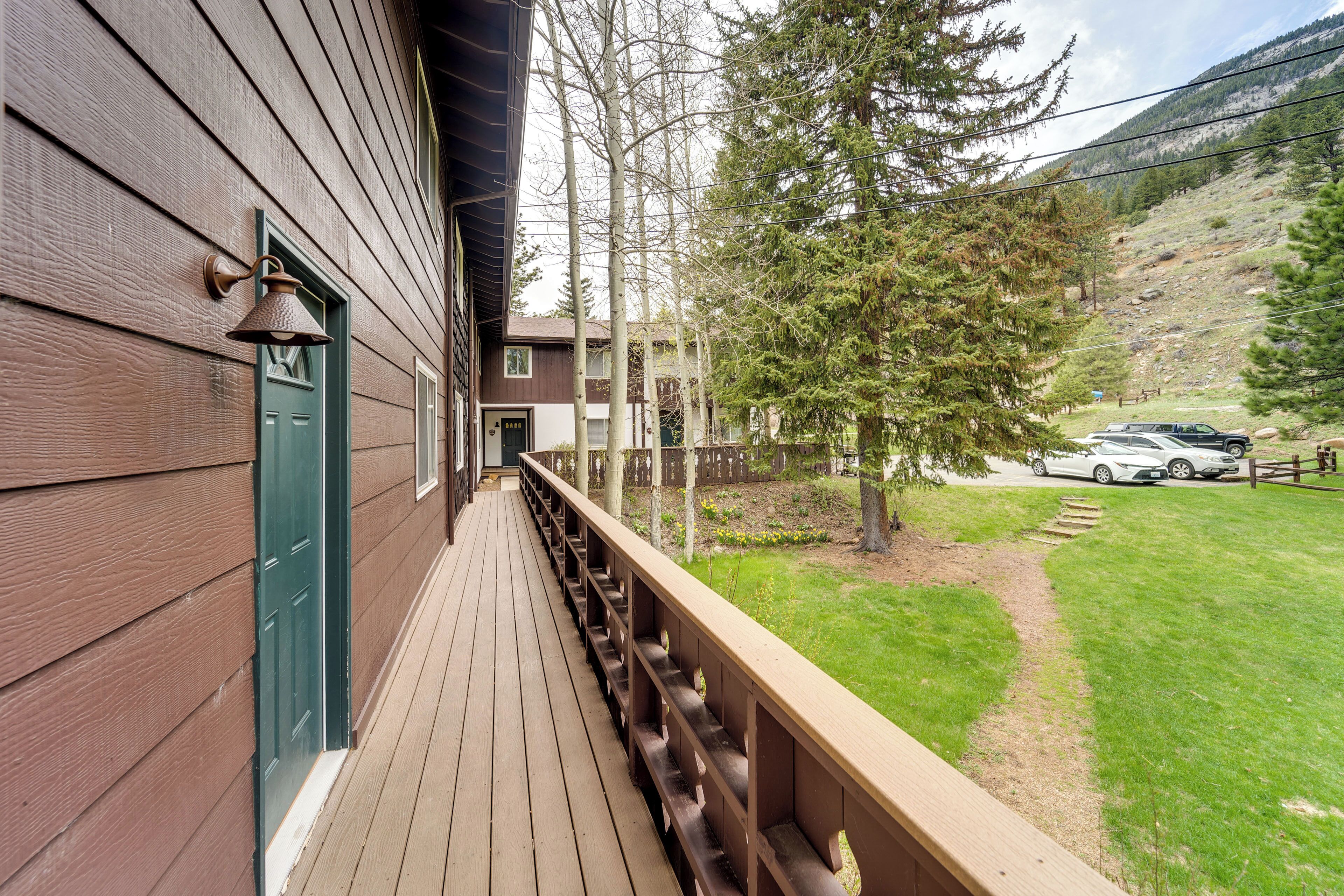 A wooden deck with a green door, a metal bell, and a view of a mountain and a car parked in the background.