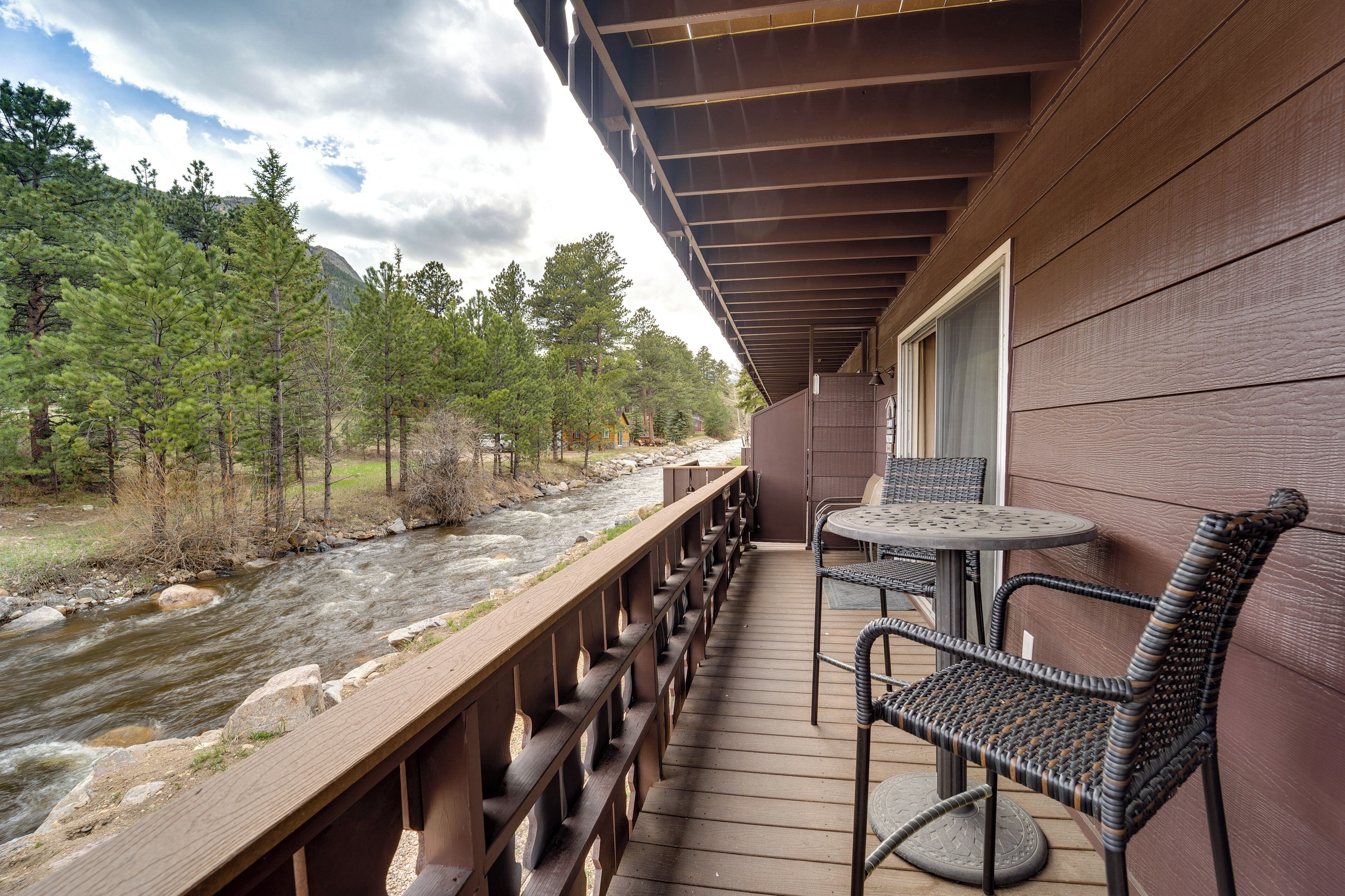 Una terraza de madera con vista a un río, mobiliario exterior y una montaña al fondo.