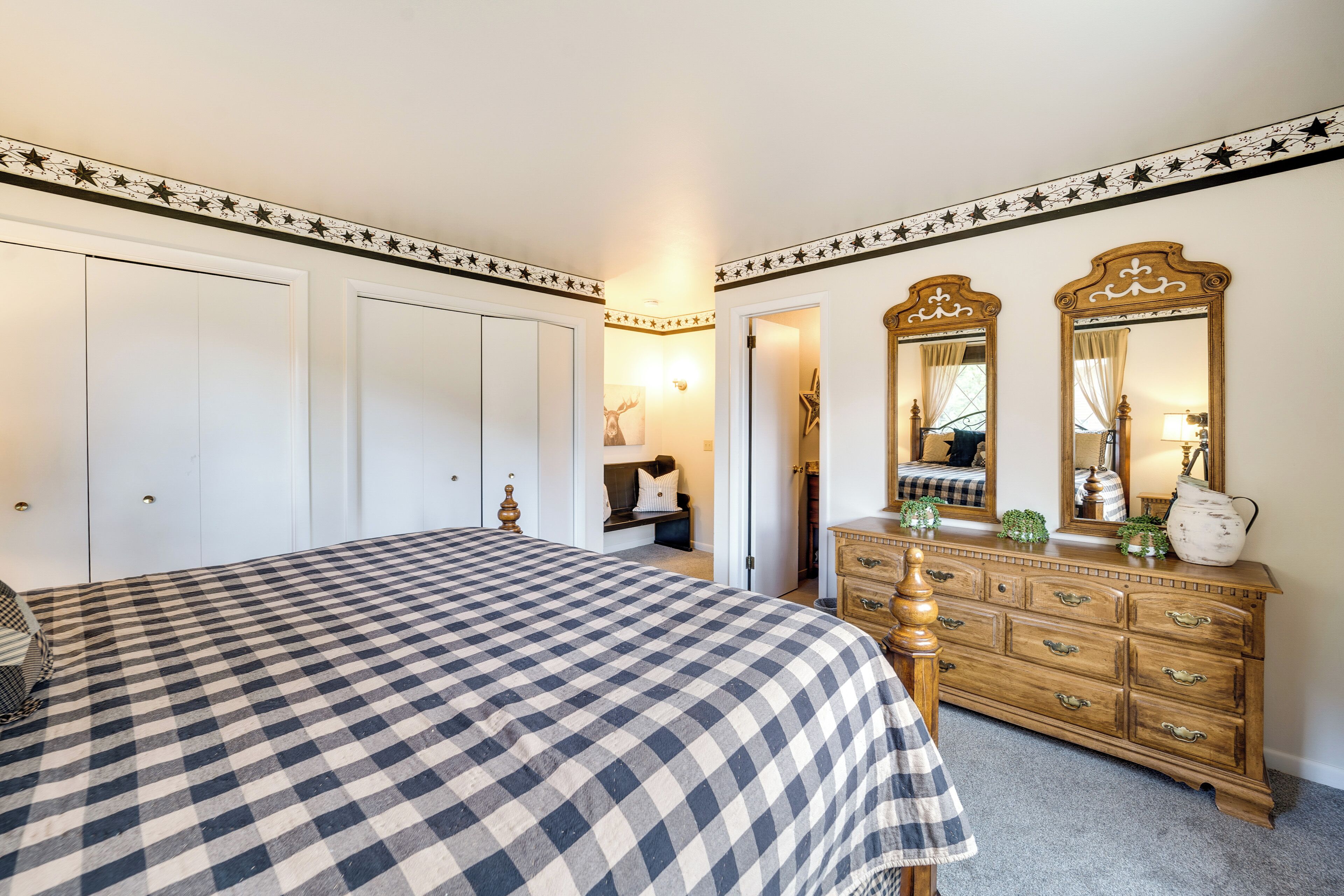 A bedroom with a checkered bedspread, a wooden dresser with drawers, a mirror, and a door leading to another room.
