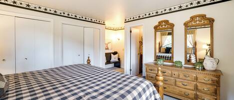 A bedroom with a checkered bedspread, a wooden dresser with drawers, a mirror, and a door leading to another room.