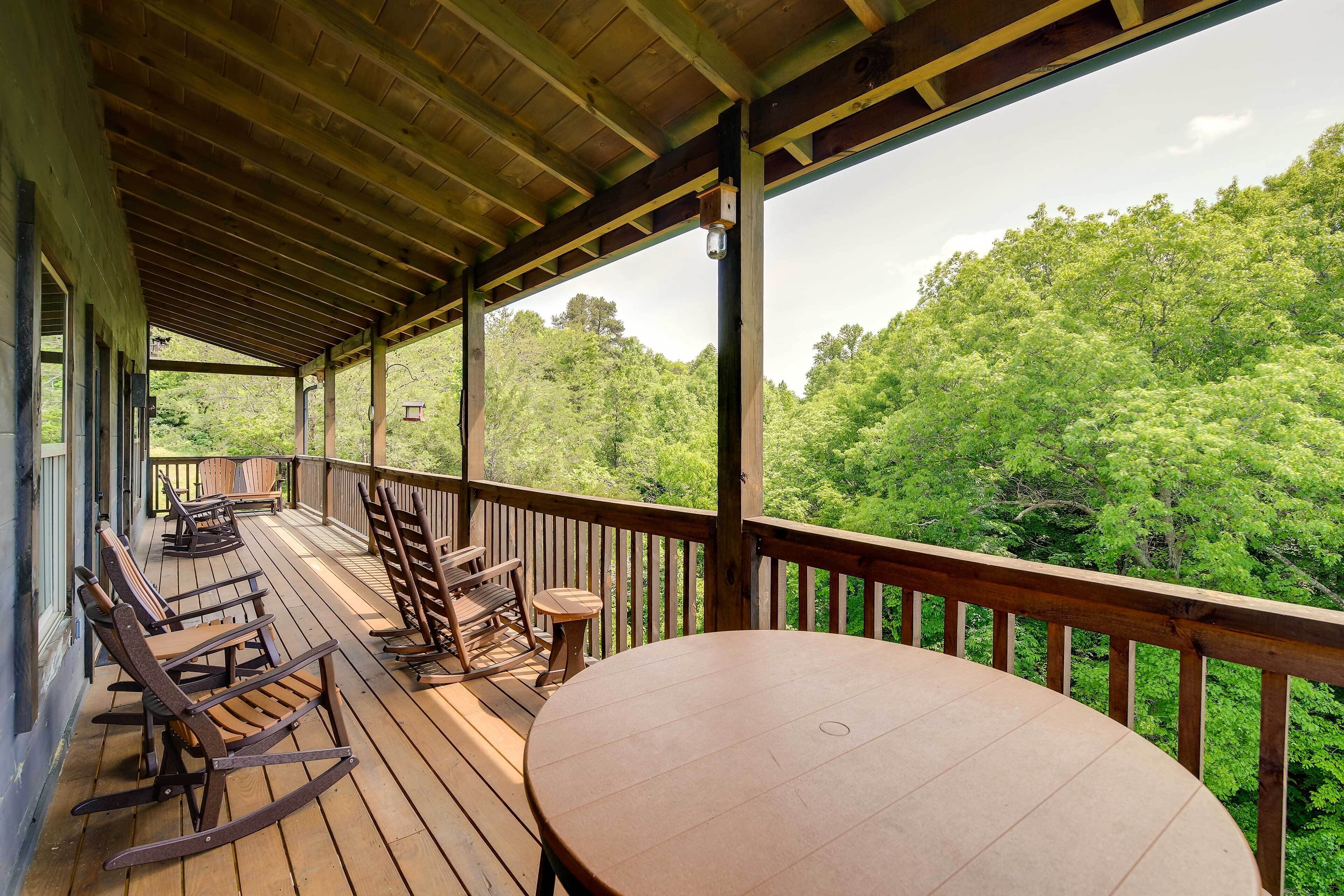 A covered outdoor deck with wooden furniture and a table, overlooking a forested area.