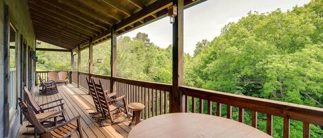 A covered outdoor deck with wooden furniture and a table, overlooking a forested area.