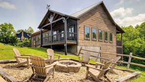 A wooden cabin with a balcony, a fire pit, and Adirondack chairs.