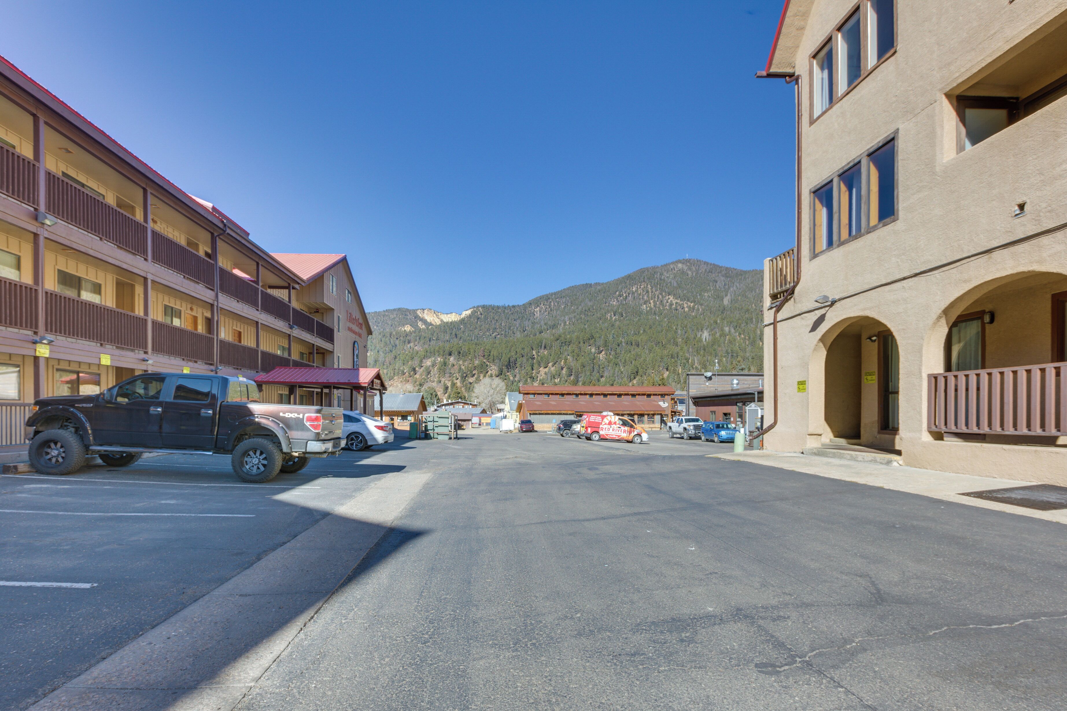 Un hotel de varios pisos con balcones, estacionamiento y montañas al fondo.
